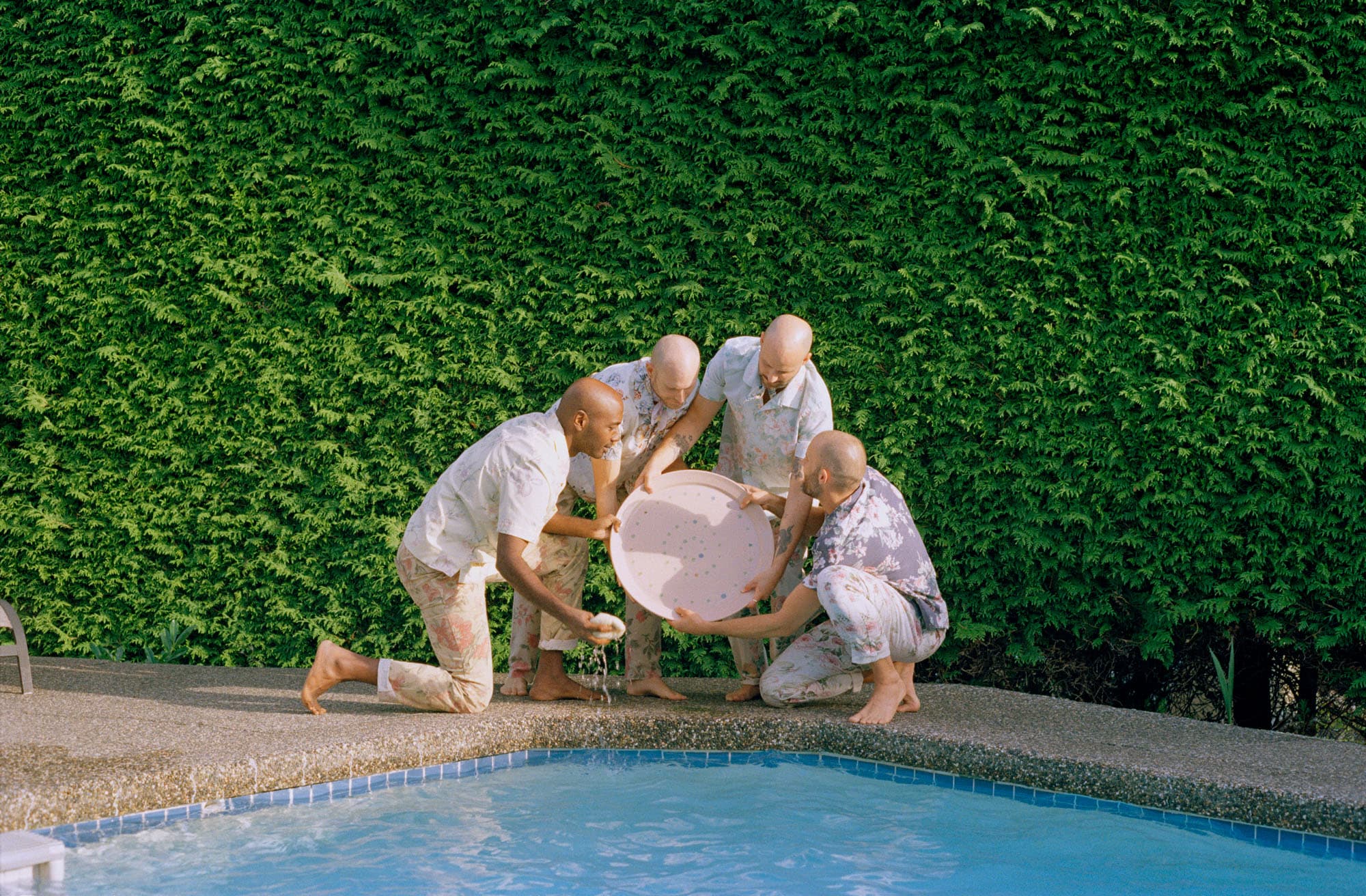 A group of four men, all bald, are gathered around a circular, light-colored tray near the edge of a swimming pool. They are dressed in floral patterned shirts and pants, with one man holding a small object. Behind them is a lush, green hedge, and the pool water is visible at the bottom of the image. The setting suggests a warm, sunny day.