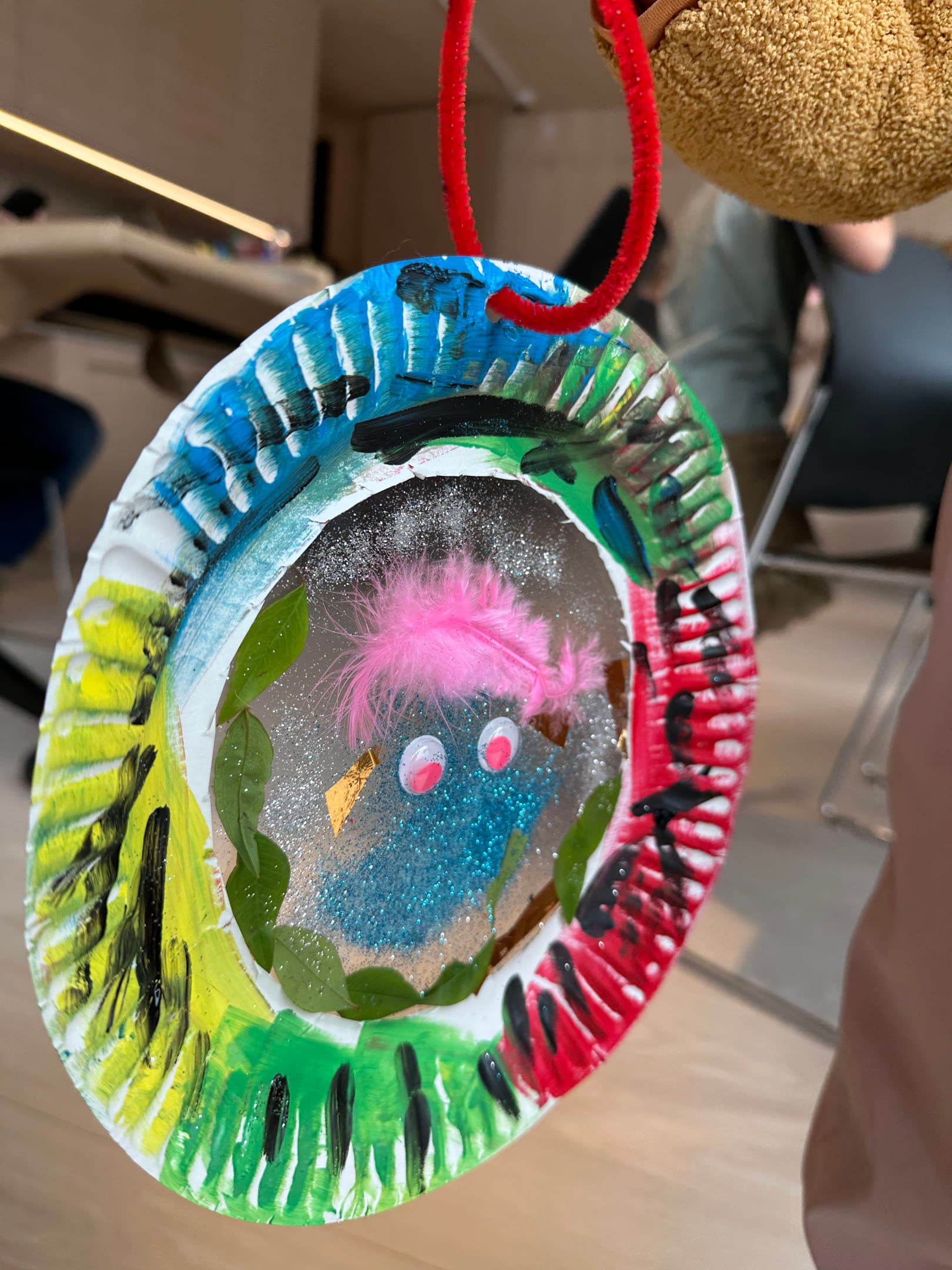 A colorful, handcrafted decoration made from a paper plate, featuring vibrant paint in green, blue, red, and black around the edges. The center contains a sparkly blue figure with a pink pom-pom for hair, surrounded by green leaves. A red pipe cleaner serves as a hanging loop at the top. In the background, a glimpse of a workshop setting is visible, with blurred figures sitting at tables.