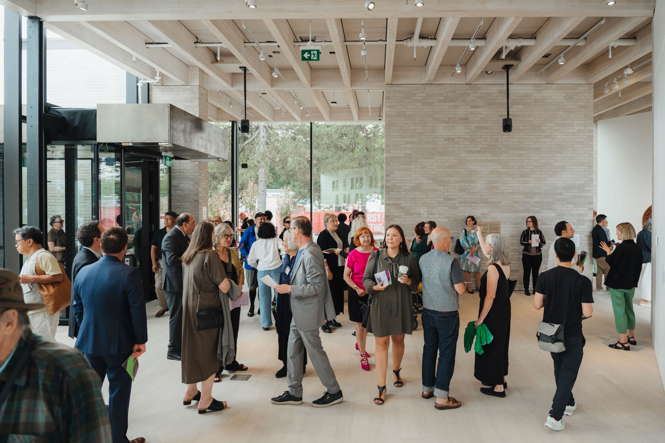 A crowded indoor space with large windows and a modern design, filled with people engaging in conversation. Attendees of various ages and styles are scattered throughout the room, some holding pamphlets. The walls are light-colored with exposed beams on the ceiling, and greenery is visible outside the windows.