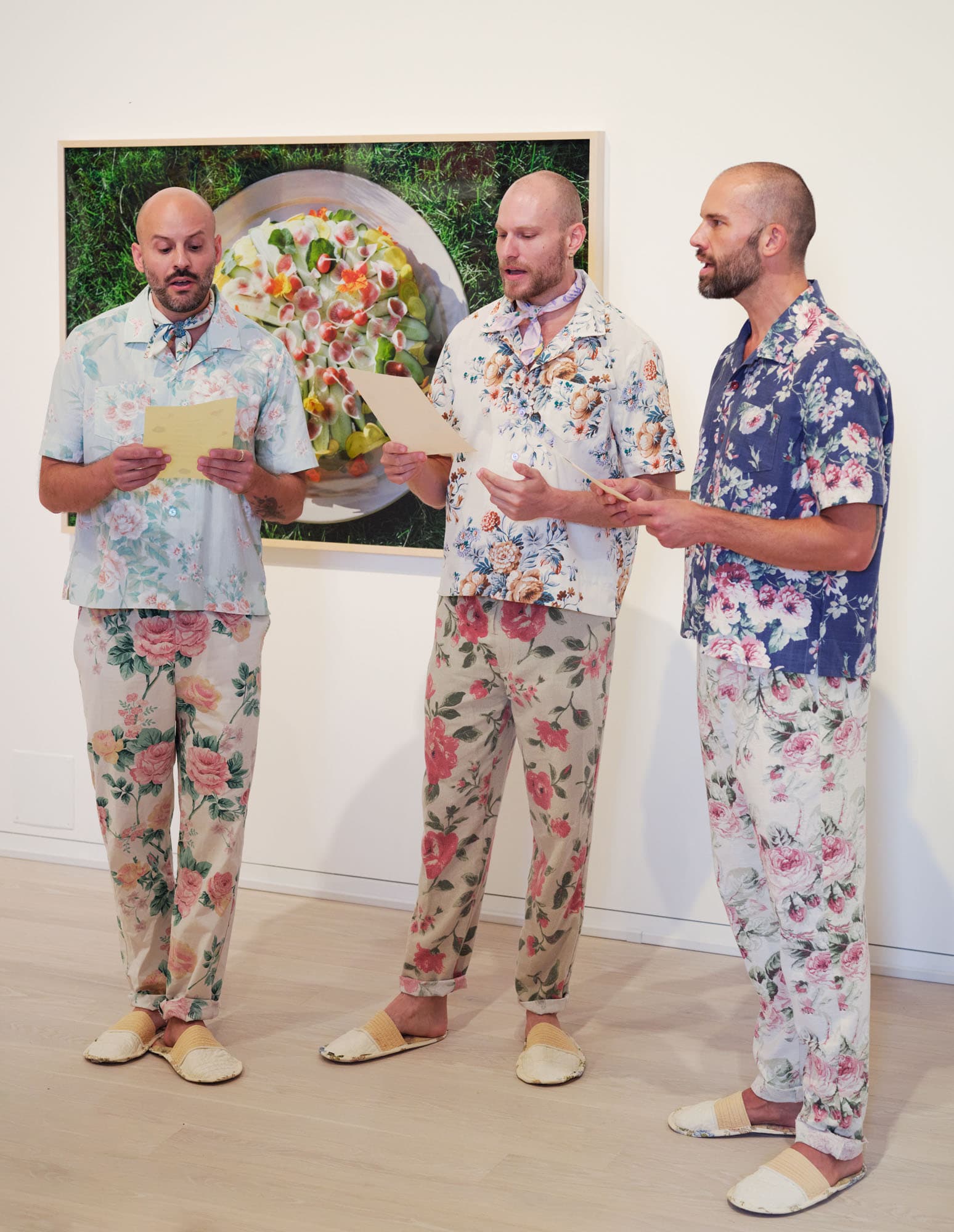 Three bald men are standing together in a gallery, dressed in matching floral-patterned shirts and pants. They each hold sheets of paper, appearing to sing or recite something. They wear comfortable slip-on shoes and are positioned in front of a large photograph of a colorful dish surrounded by greenery. The gallery has a light wood floor and white walls.