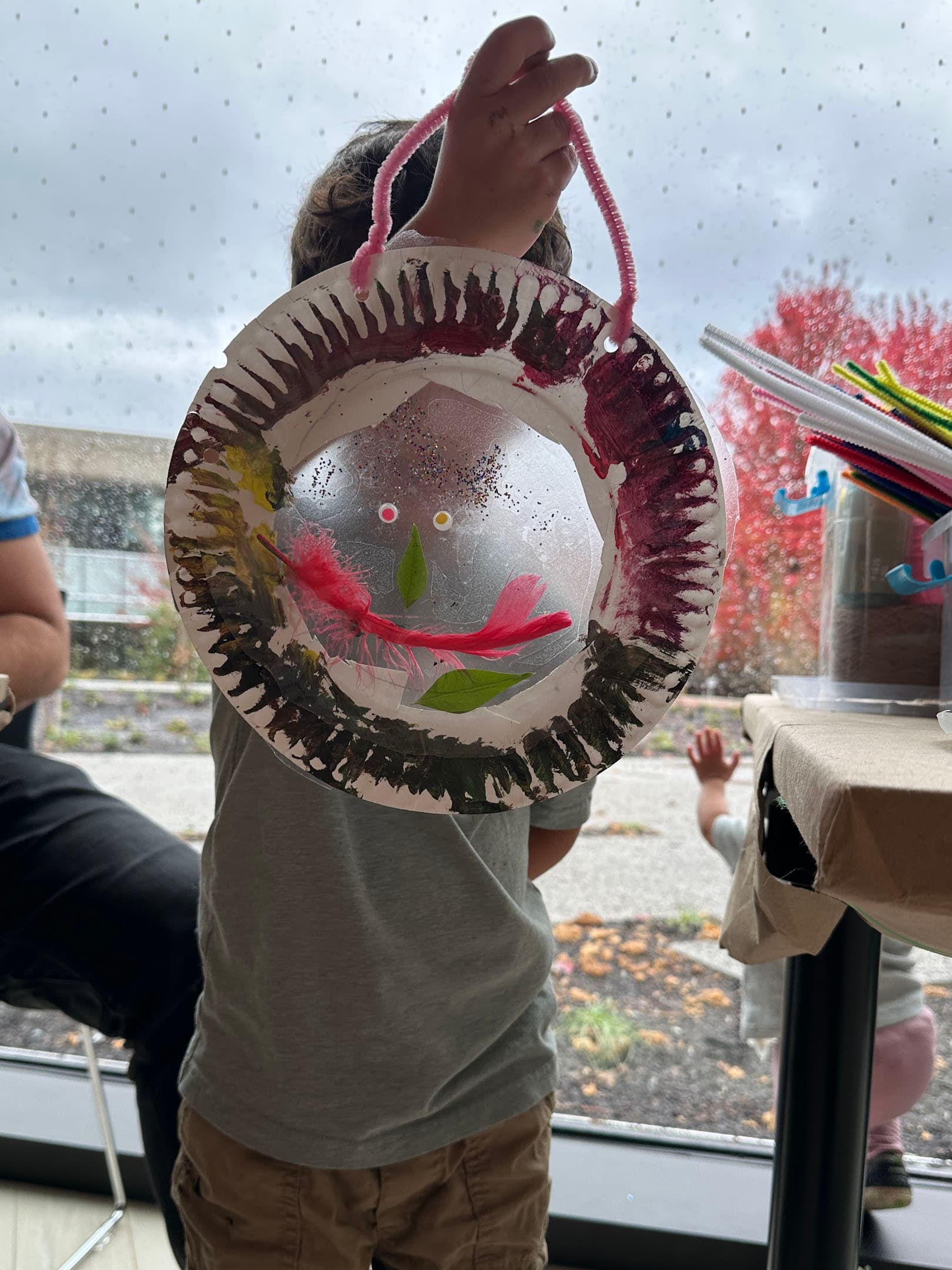 A child holds up a colorful, circular art piece made from paper plates, featuring painted designs and decorative elements like feathers and leaves. The background shows raindrops on a window and a glimpse of an outdoor scene with colorful foliage. Other children can be seen in the background, one waving from outside.