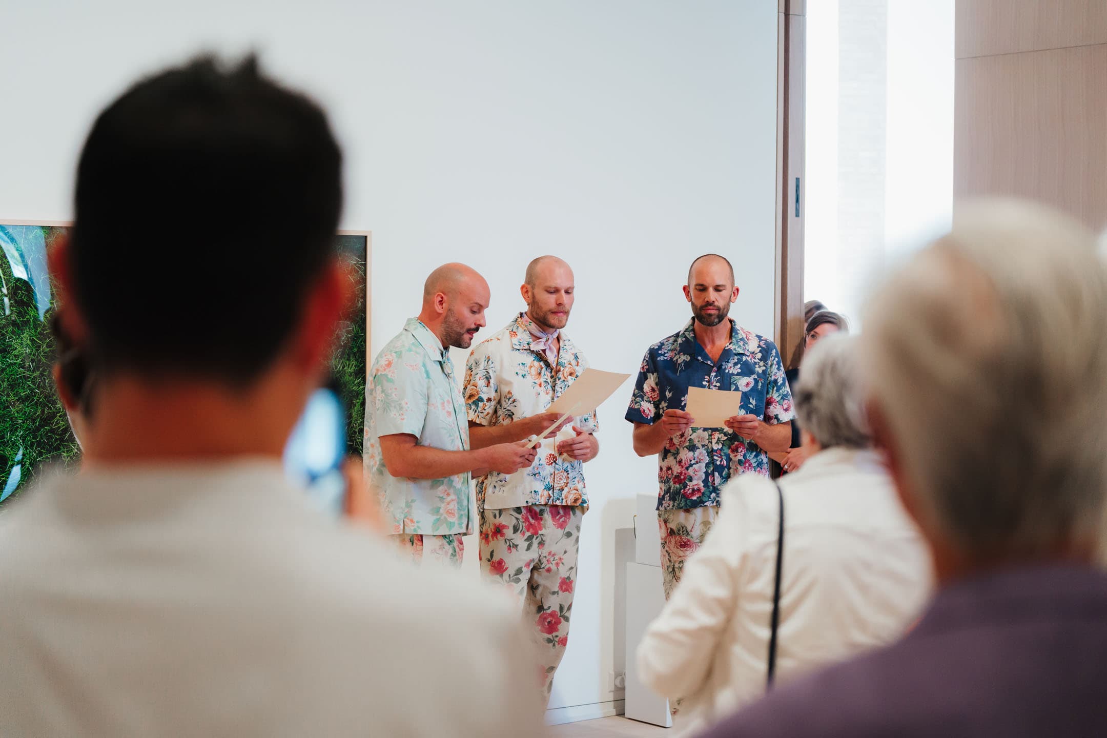 Three men in floral-patterned outfits stand in front of an audience, reading from sheets of paper. The background features a large artwork, while several people are visible in the foreground, observing the scene. The setting appears to be a gallery or event space with bright lighting.