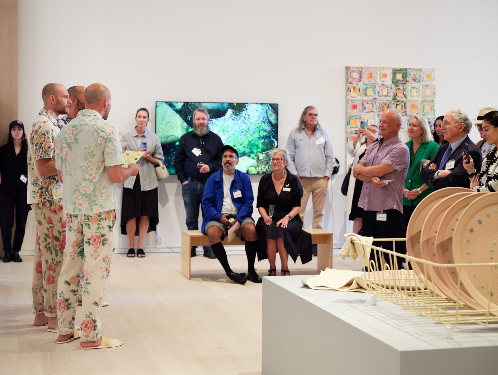 A group of four men in floral shirts and pants stand in the foreground, engaging with an audience in an art gallery setting. Various attendees, including a mix of men and women, observe and listen intently. In the background, there is a large screen displaying images of nature, alongside a colorful artwork on the adjacent wall. A table with sculptural art pieces is visible in the foreground, adding to the exhibit's ambiance. The gallery features light wood flooring and a bright, spacious atmosphere.