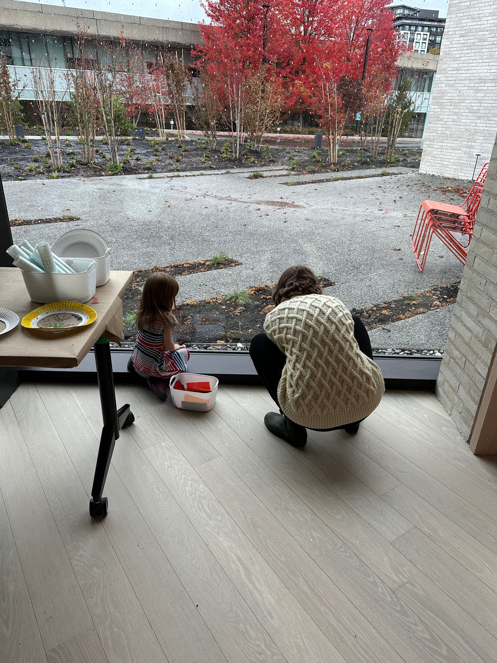 A woman and a young girl sit on a light wooden floor next to a large window. The woman, wearing a cream-colored knitted sweater and black pants, is crouching beside the girl, who is wearing a striped dress. They are both looking outside at a landscaped area with colorful autumn trees and a path. There are plates and utensils on a table nearby.