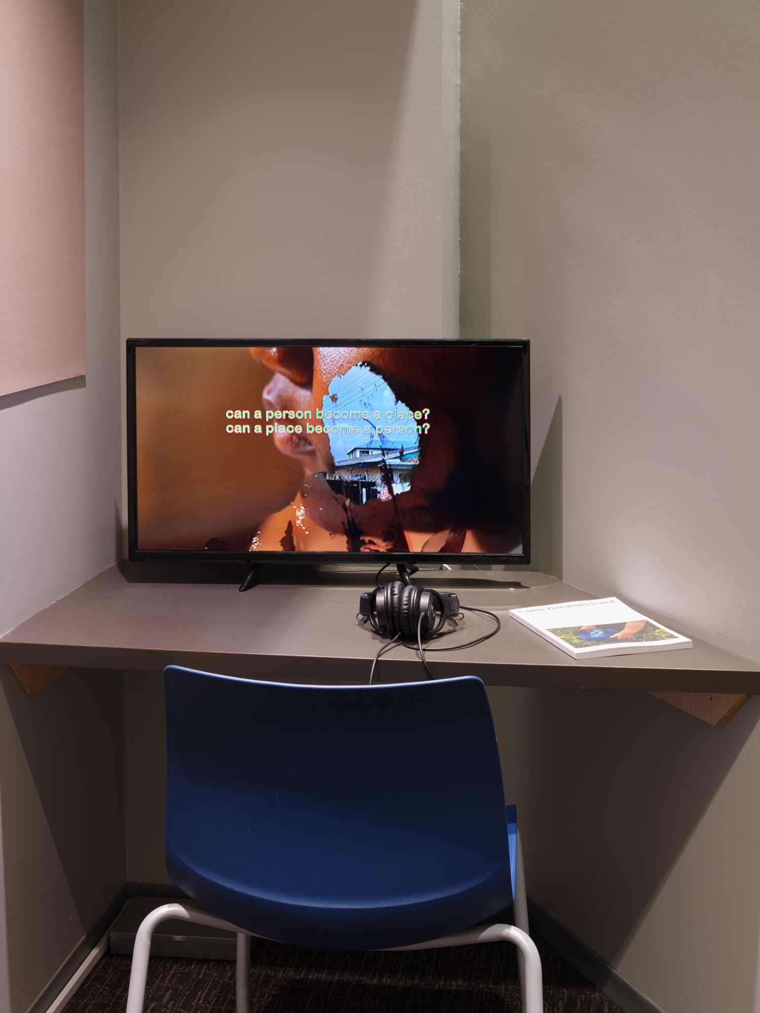 A desk with a dark gray surface features a television displaying text that asks philosophical questions about identity and place. A pair of black headphones is placed on the desk beside the monitor. A blue chair sits in front of the desk, and a book is visible on the surface. The wall behind is a muted gray, with a light pink area visible to the left.