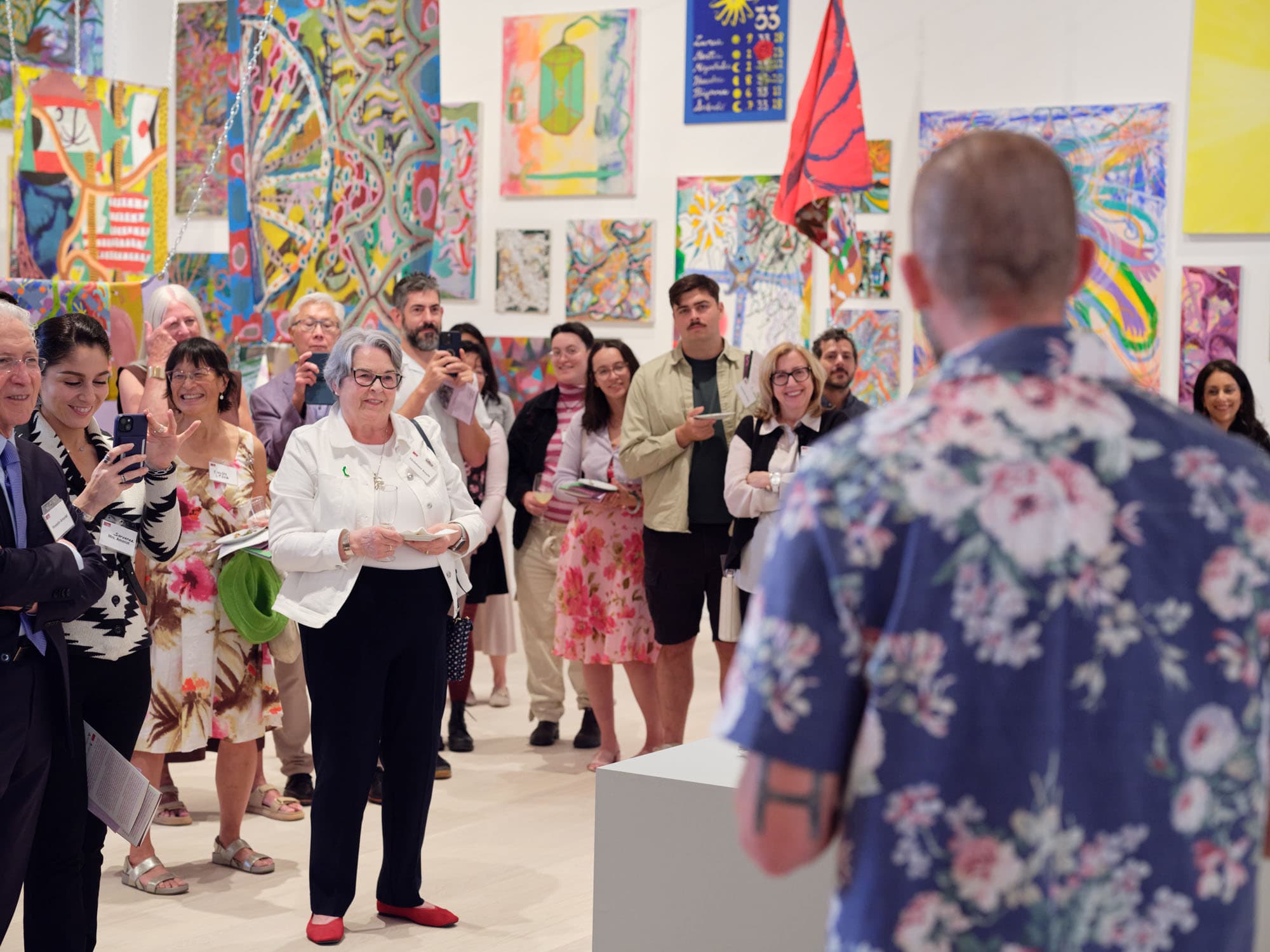 A crowd of people gathers in an art gallery, with colorful abstract paintings displayed prominently on the walls. In the foreground, a speaker in a floral shirt addresses the audience, while several attendees hold mobile phones and notebooks, engaged in the presentation. Some individuals appear to be smiling or taking notes, contributing to a lively atmosphere.