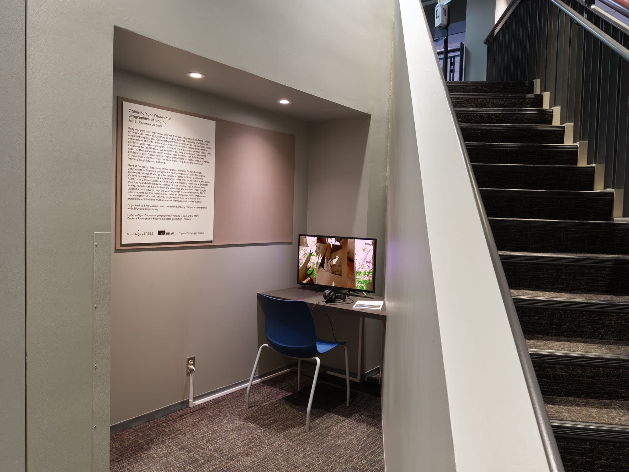 A small workspace is shown with a blue chair and a desk featuring a monitor displaying a video. On the wall behind the desk is an informational poster about an exhibit titled "Ophthalmologist Obsessions: Spectacles of Longing." A staircase is visible on the right, leading to an upper level. The area is well-lit with spotlights above the desk.