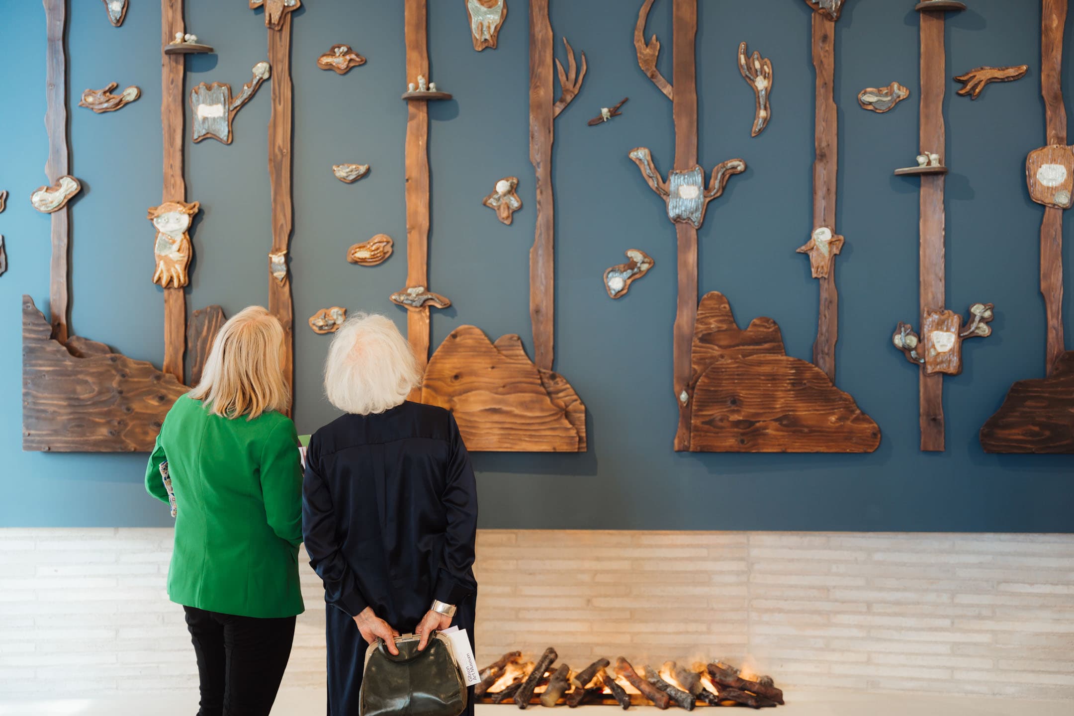 Two women stand facing a large art installation on a blue wall, which features wooden tree-like structures adorned with ceramic pieces resembling animals and natural elements. One woman wears a green blazer and holds a magazine, while the other, with white hair, carries a bag. A modern fireplace with logs is visible at the bottom of the wall.