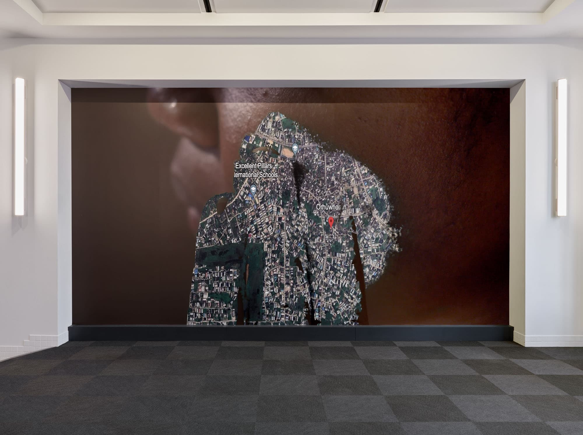 A large mural in a room features an aerial view of a neighborhood overlaid on a close-up of a person's shoulder and neck. The mural includes labels for "Excellent Pillars International Schools" and a location marked as "Orhuwhor." The room has gray carpet with a checkered pattern, and bright wall-mounted lights on either side of the mural.
