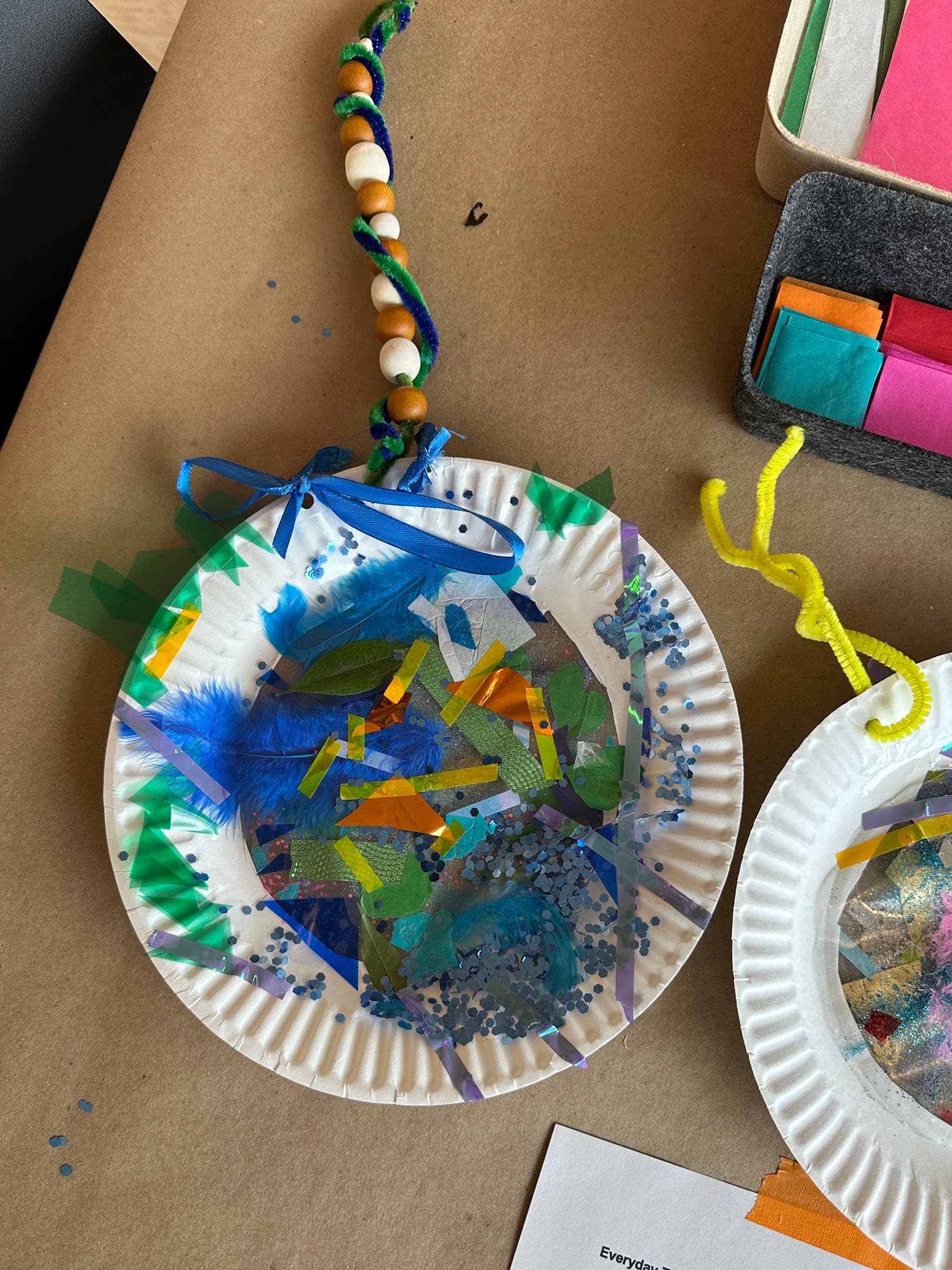 A decorated paper plate hangs from colorful beads and pipe cleaners. The plate is adorned with blue feathers, various colored strips of paper, shiny confetti, and green shapes. Nearby, there are additional craft materials, including colored tissue paper, on a brown surface.