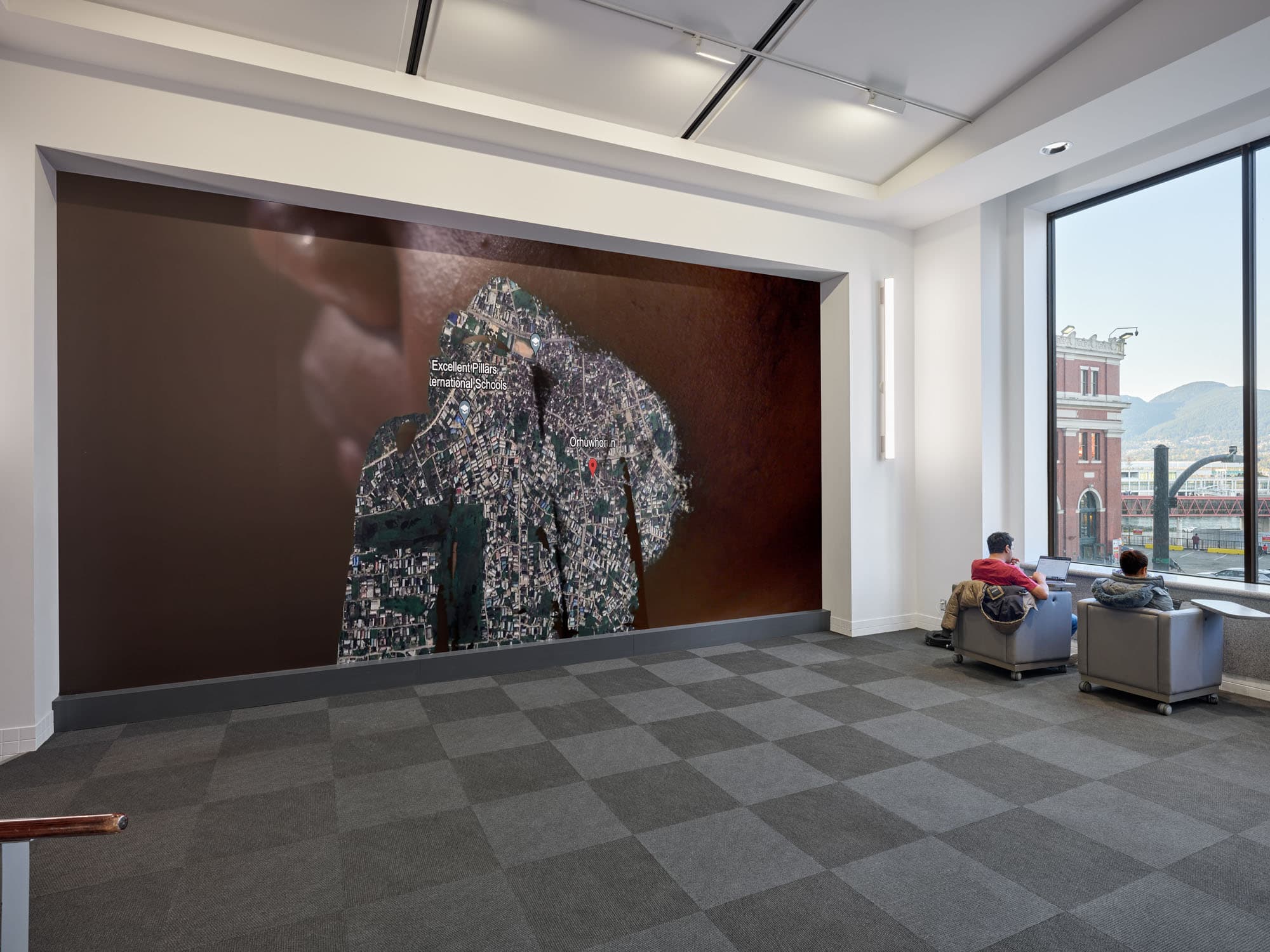 A spacious interior featuring a large wall mural of an aerial view of a city, with labeled landmarks. Two individuals sit in gray armchairs against a window, looking out at a view of a red brick building and distant mountains. The floor has a checkered gray carpet pattern, and soft lighting illuminates the room.