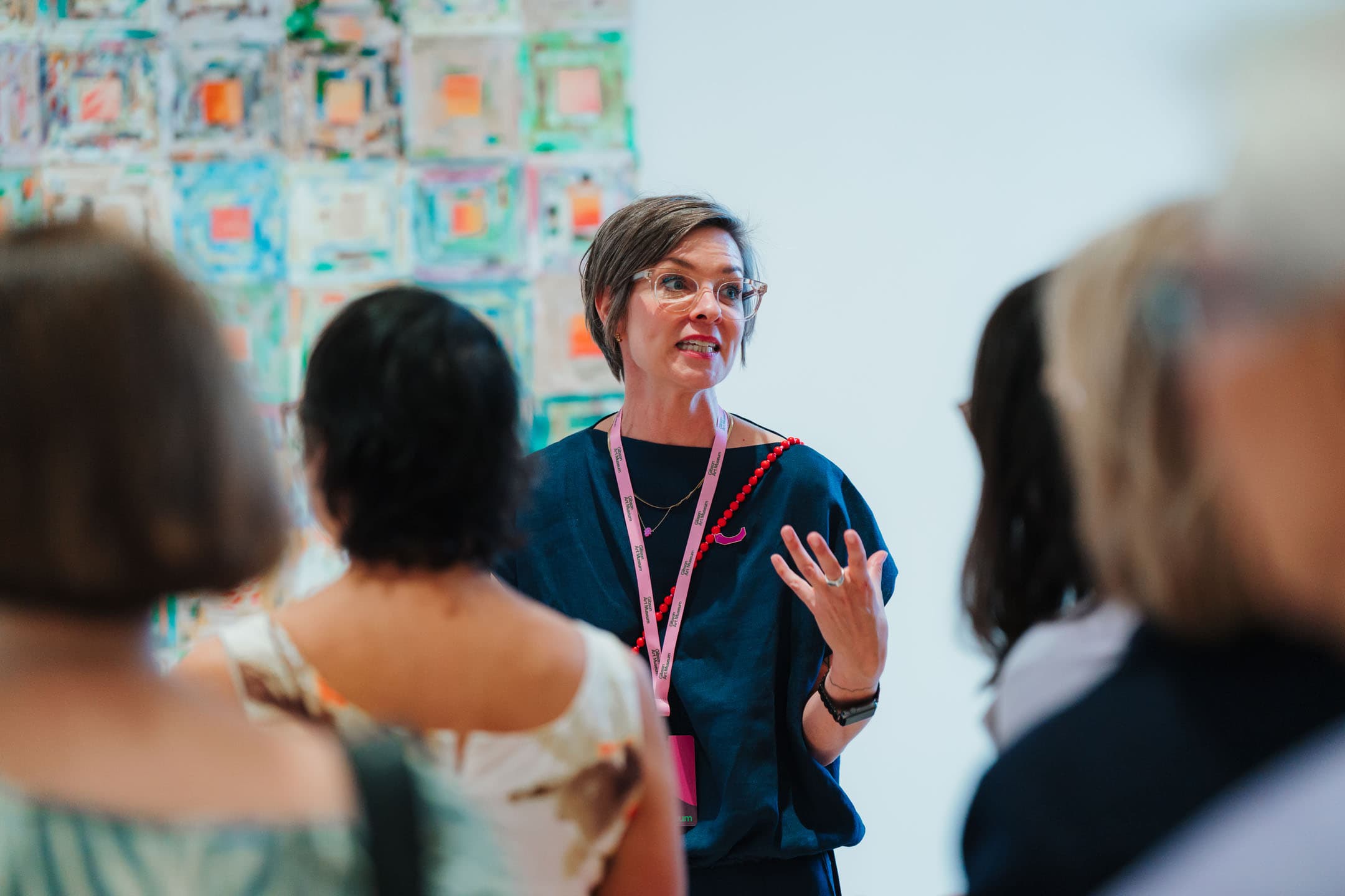 A woman with short hair and glasses gestures expressively while speaking to a group of people. She is wearing a blue top and a pink lanyard. In the background, there is an abstract wall art featuring colorful geometric patterns. The audience is partially visible, with their backs facing the camera.