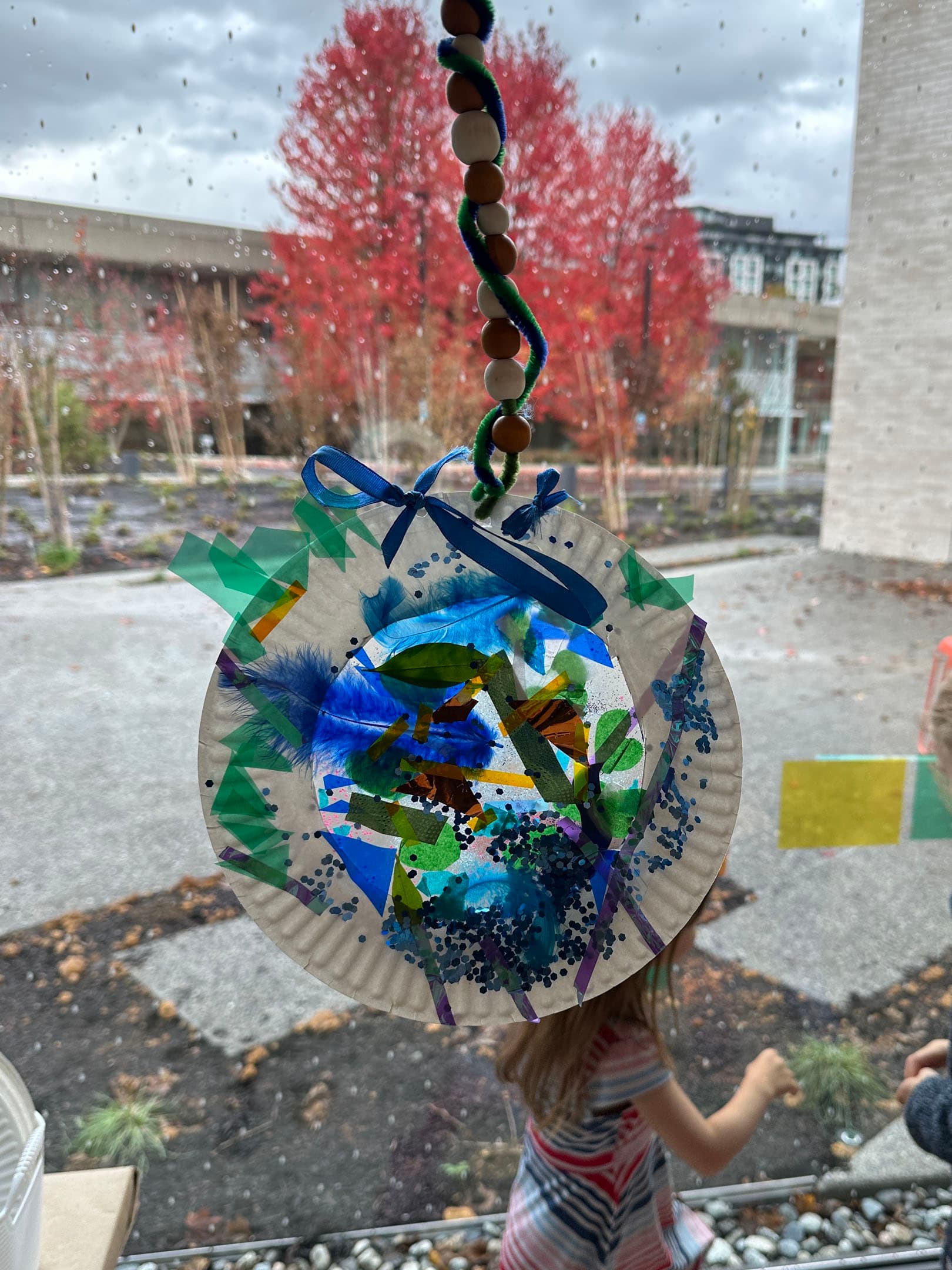 A colorful paper plate craft adorned with blue ribbons, feathers, and various green and blue decorations hangs in front of a window. Outside, there are bright red trees and a cloudy sky, with raindrops visible on the glass. In the foreground, a child's arm and part of their body can be seen, suggesting they are playing nearby.