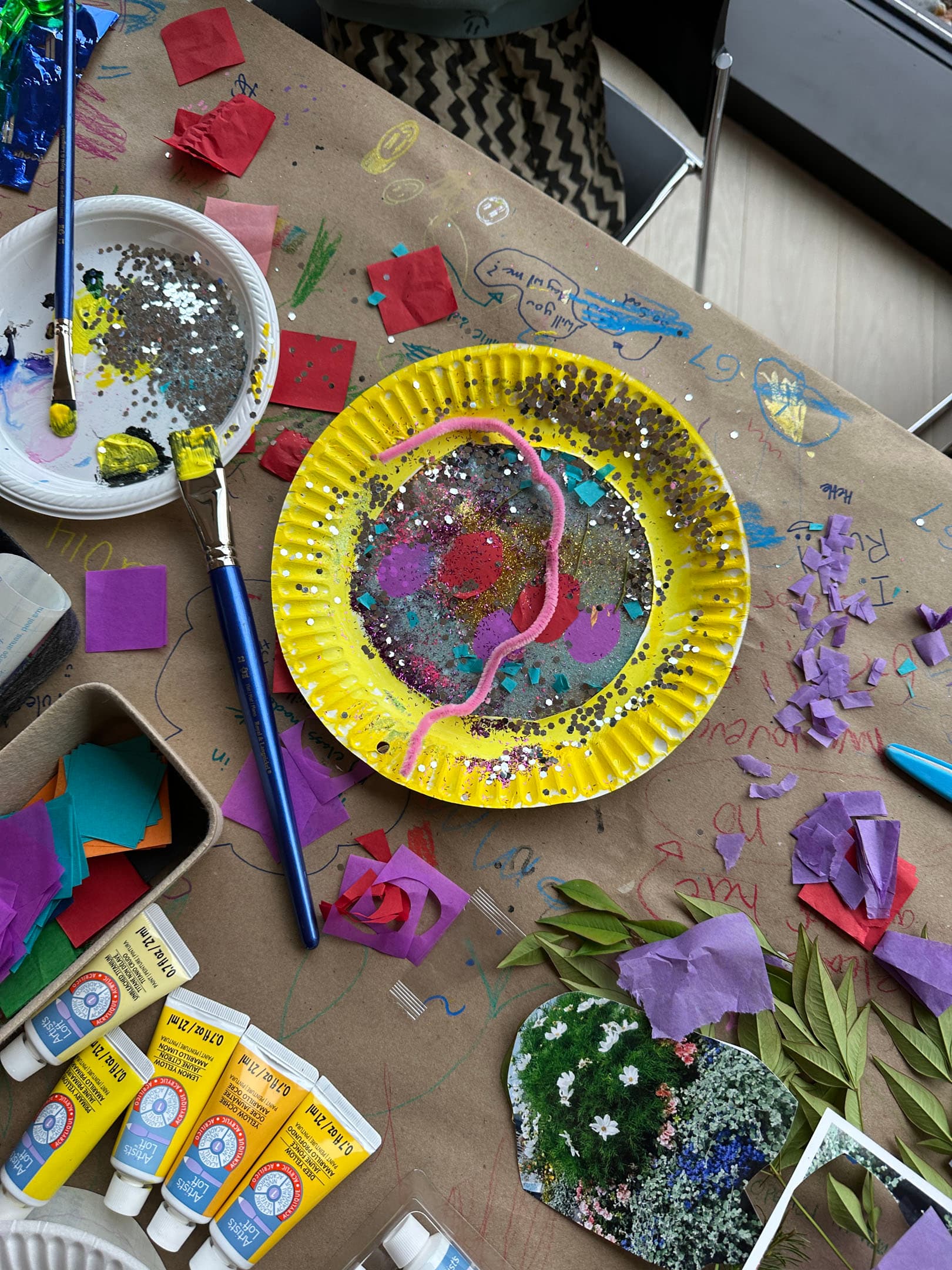 An overhead view of a creative workspace featuring a yellow paper plate decorated with colorful paint, glitter, and a pink swirl. Surrounding it are various craft materials including scattered pieces of colored paper, paintbrushes, paint tubes, and a small collage of leaves and flowers. The table is covered with brown craft paper, showing scribbles and additional colorful cutouts.
