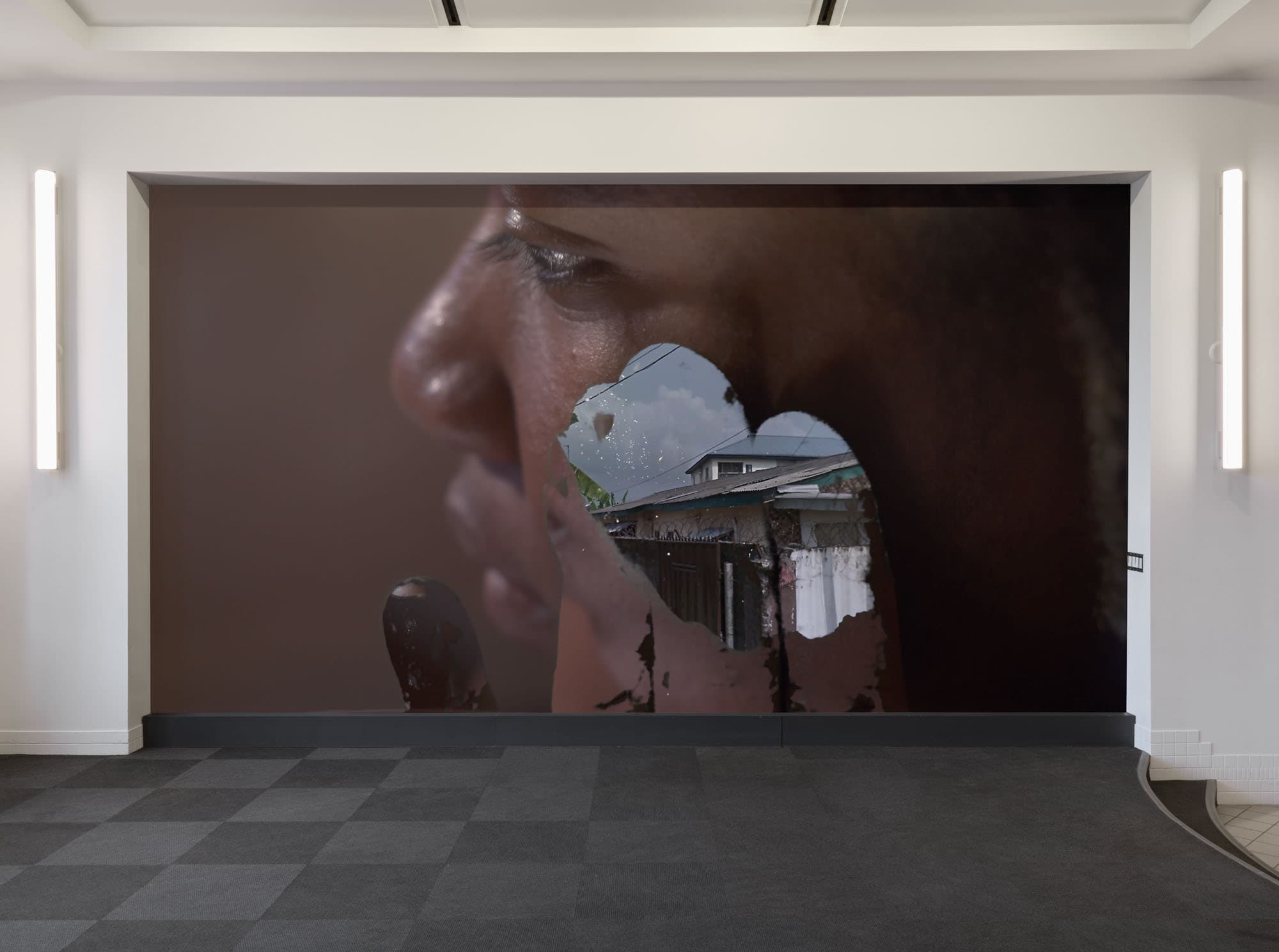 A large mural featuring a close-up of a person's profile is displayed on a wall. The individual's face seems to be overlaid with an image of a cloudy sky and a building with a white curtain, creating a layered effect. The surrounding space has a modern, minimalist design with gray flooring and wall-mounted lights.