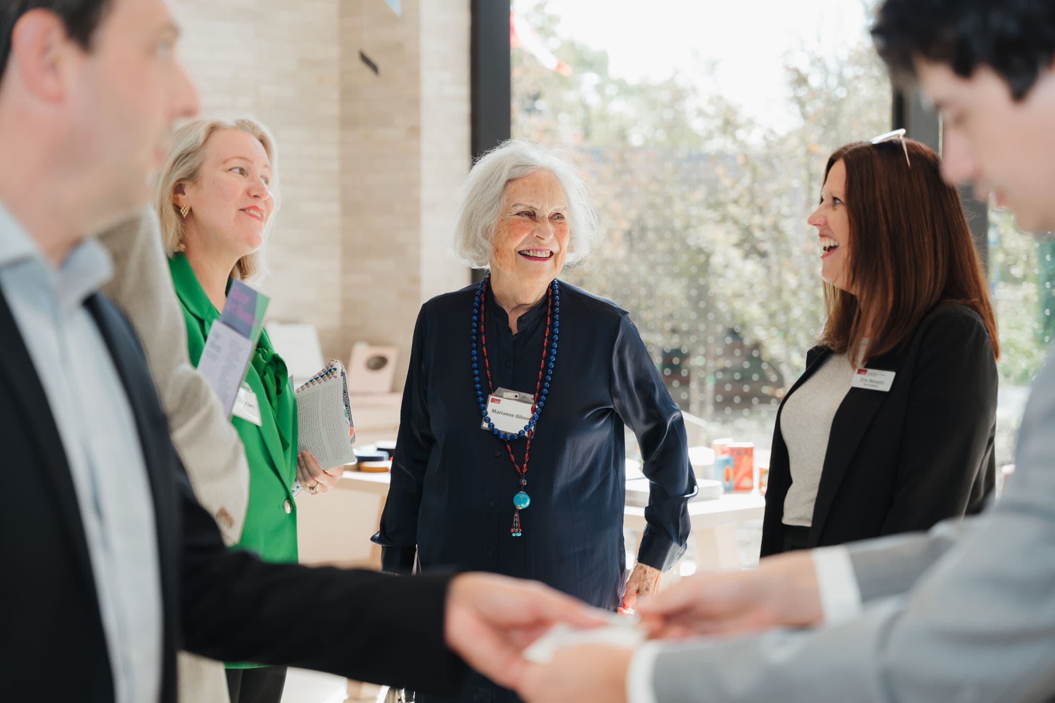 A group of four people are engaged in conversation in a bright, modern indoor setting. One older woman with gray hair, dressed in a navy blouse and colorful necklace, smiles as she speaks. A middle-aged woman in a green blazer holds a notepad and appears to be listening intently. Two younger individuals, one in a black blazer and the other in a light gray suit, are partially visible and seem to be exchanging business cards. The background features soft sunlight filtering through large windows and a glimpse of greenery outside.