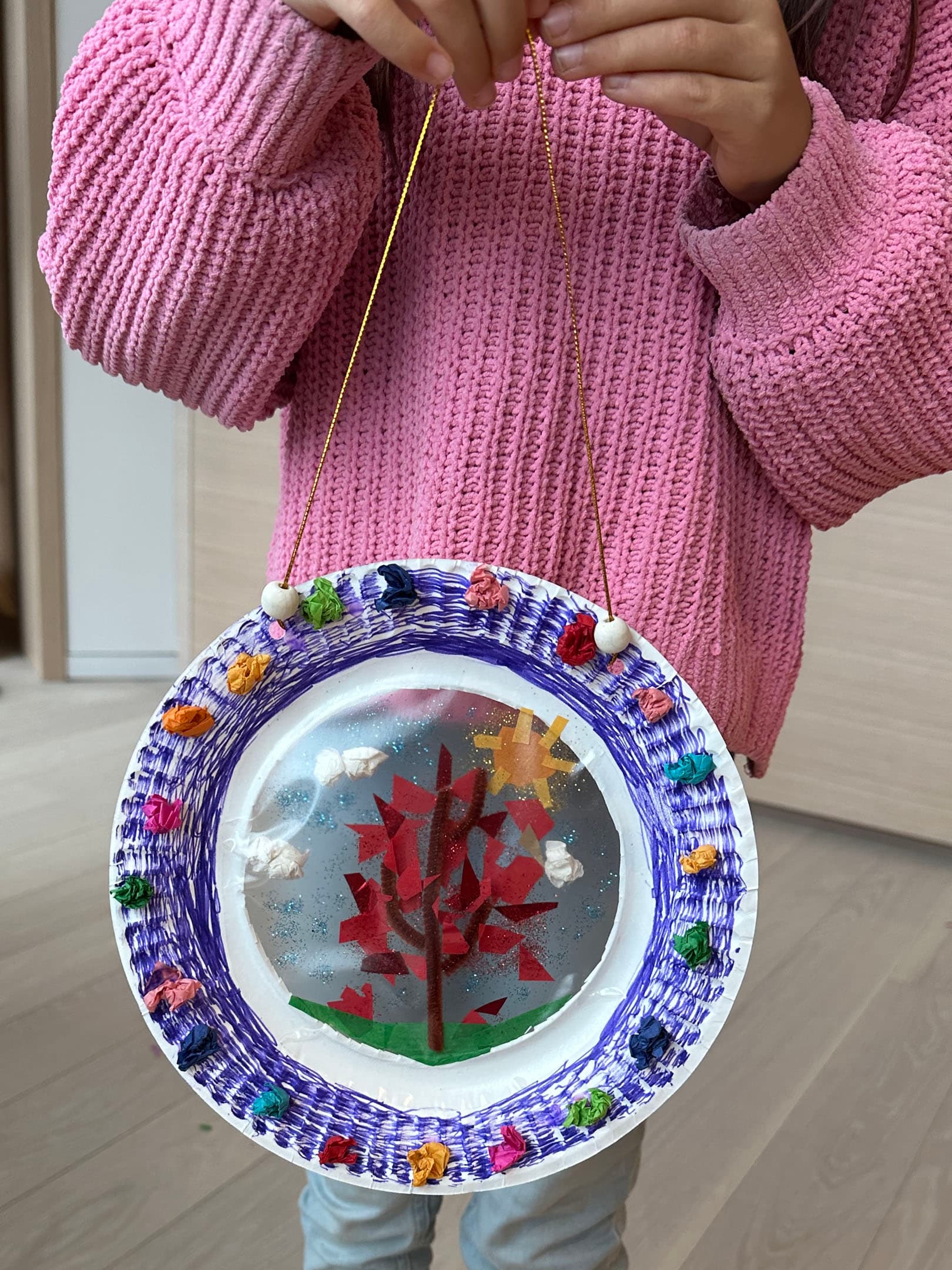 A child wearing a pink sweater holds a circular craft project decorated with colorful bits of paper and a golden string for hanging. The artwork features a red tree with a bright sun and clouds, encapsulated in transparent material. The background is a light wooden floor, adding a warm ambiance to the scene.
