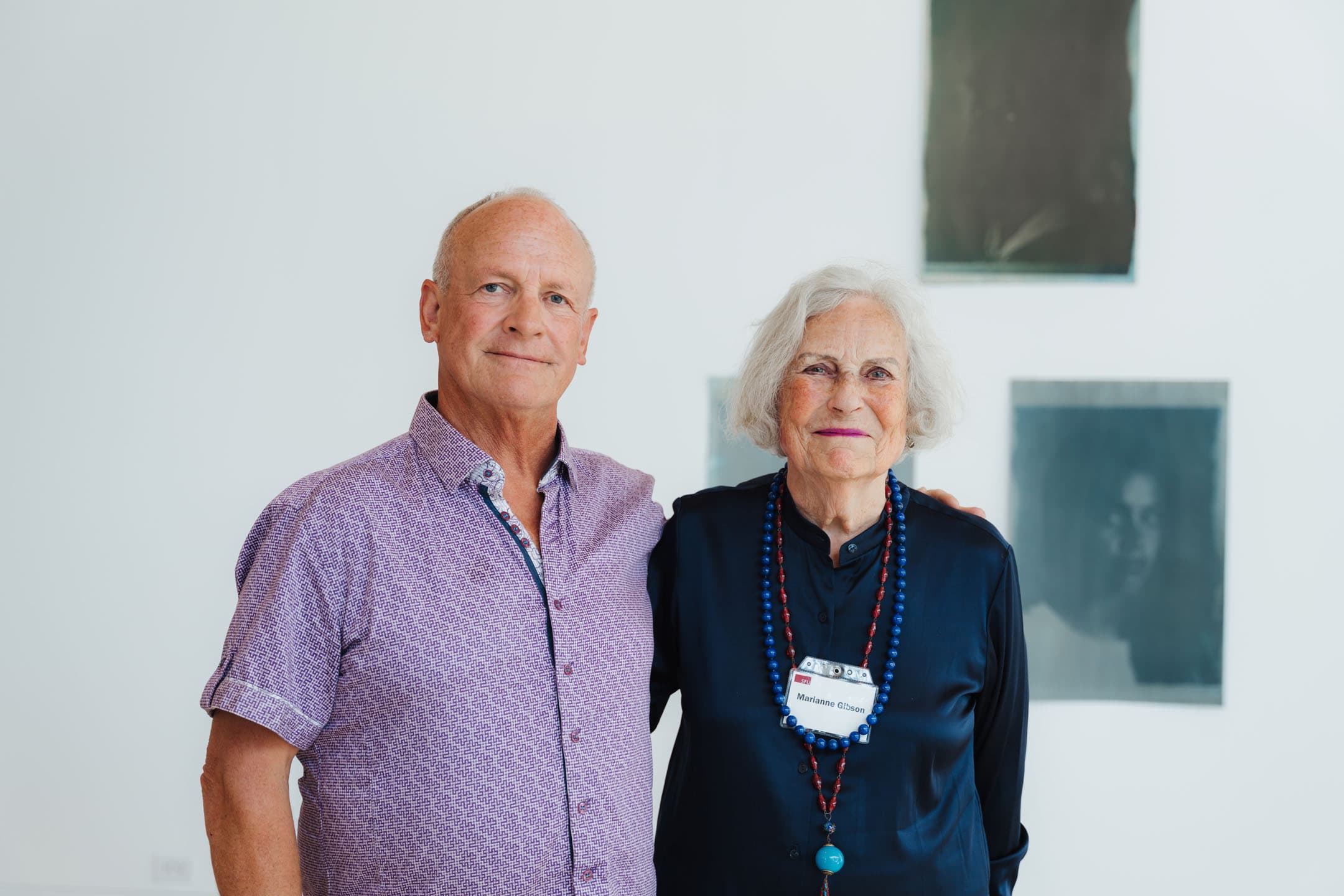 A man and a woman stand close together, smiling at the camera against a light-colored wall. The man, wearing a short-sleeved, patterned shirt, has a serious expression. The woman, dressed in a dark blouse, has gray hair and is adorned with a necklace of colorful beads. She wears a name tag. In the background, there are several dark-toned images or artworks on the wall.