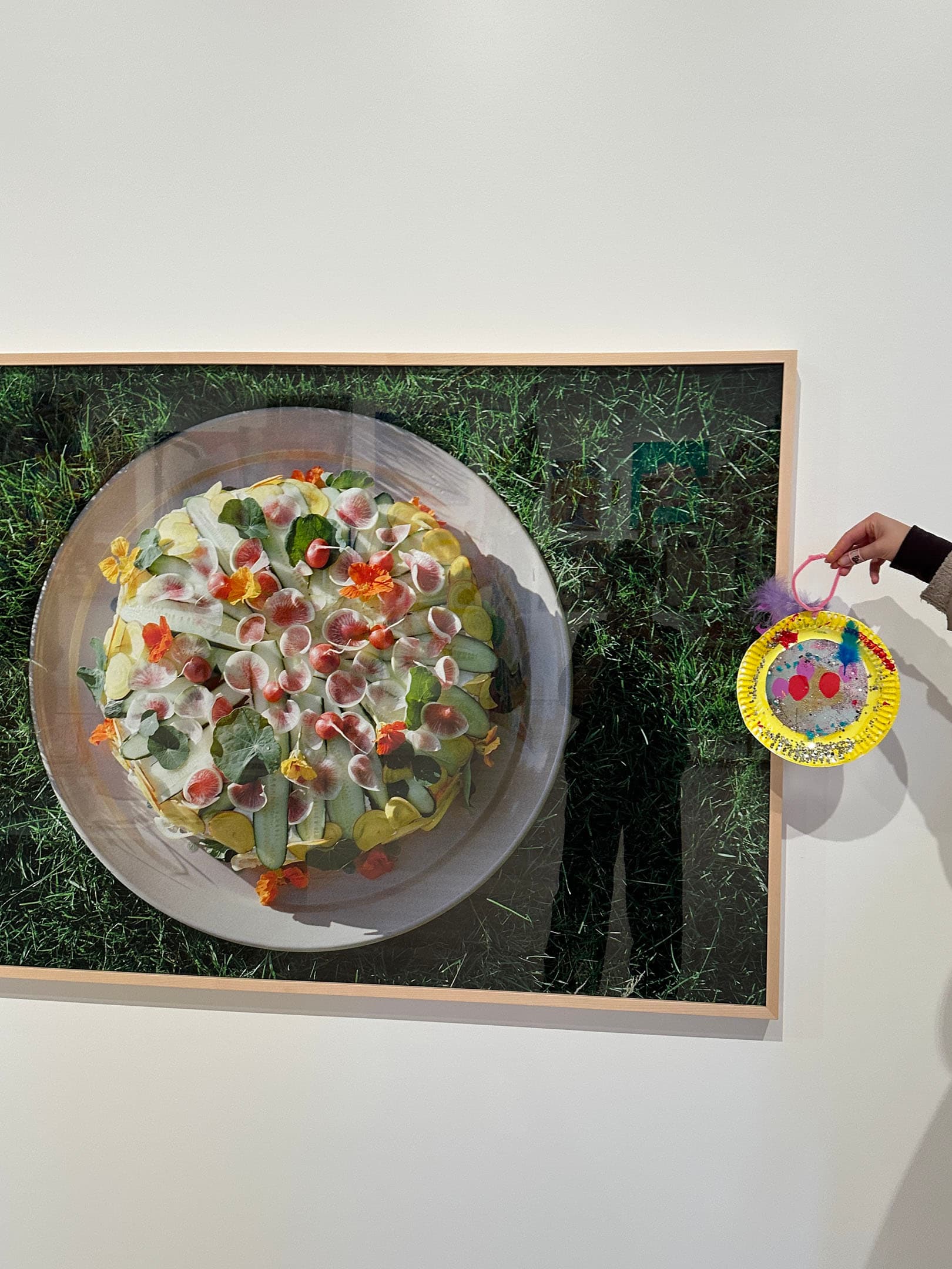 A large image of a colorful salad displayed on a shallow plate sitting on grass. The salad features various ingredients like greens, edible flowers, and sliced vegetables. To the right, a person holds a decorative plate with a painted design in bright colors, partially obscured by their hand. The background is a simple white wall.