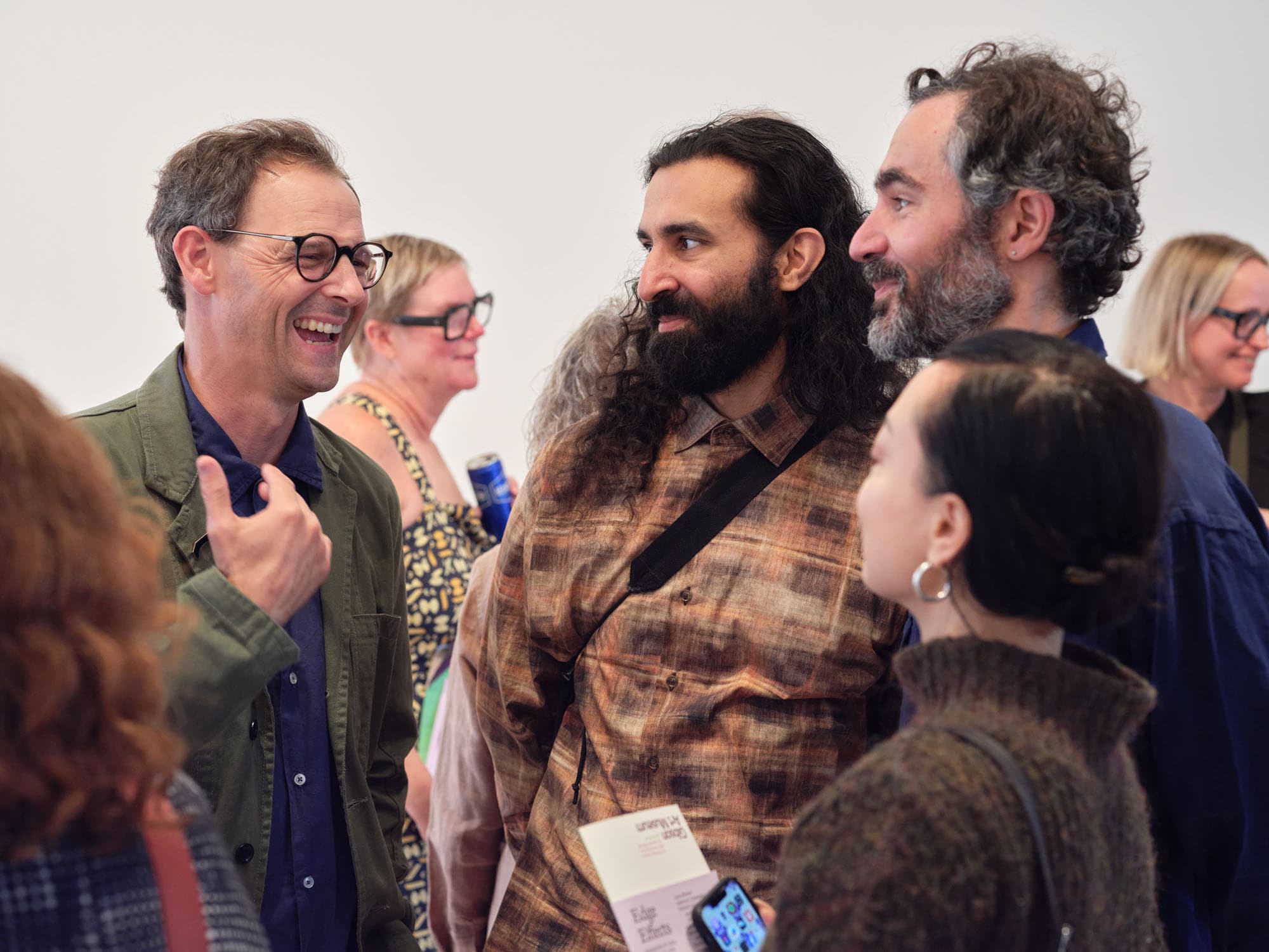 A group of four people engaged in a conversation at an event. The man on the left, wearing glasses and a green jacket, is smiling and gesturing. Beside him is a man with long curly hair and a patterned shirt. There is a woman with brown hair and earrings, looking attentively at the two men. In the background, another woman with glasses is visible, holding a can. The setting appears to be an art gallery or similar venue.