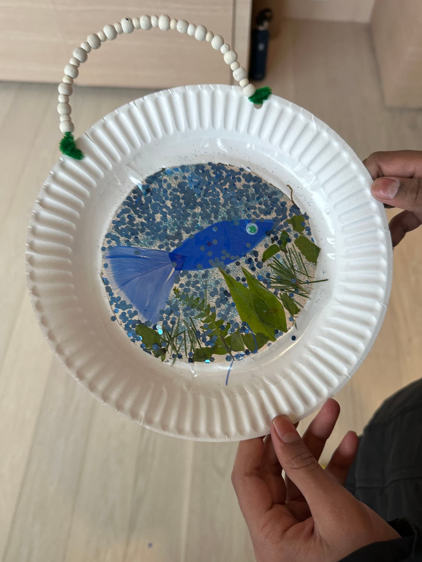 A person is holding a paper plate decorated with a blue fish, green leaves, and blue glitter. The plate has a bead and pipe cleaner handle. The background features light-colored flooring.