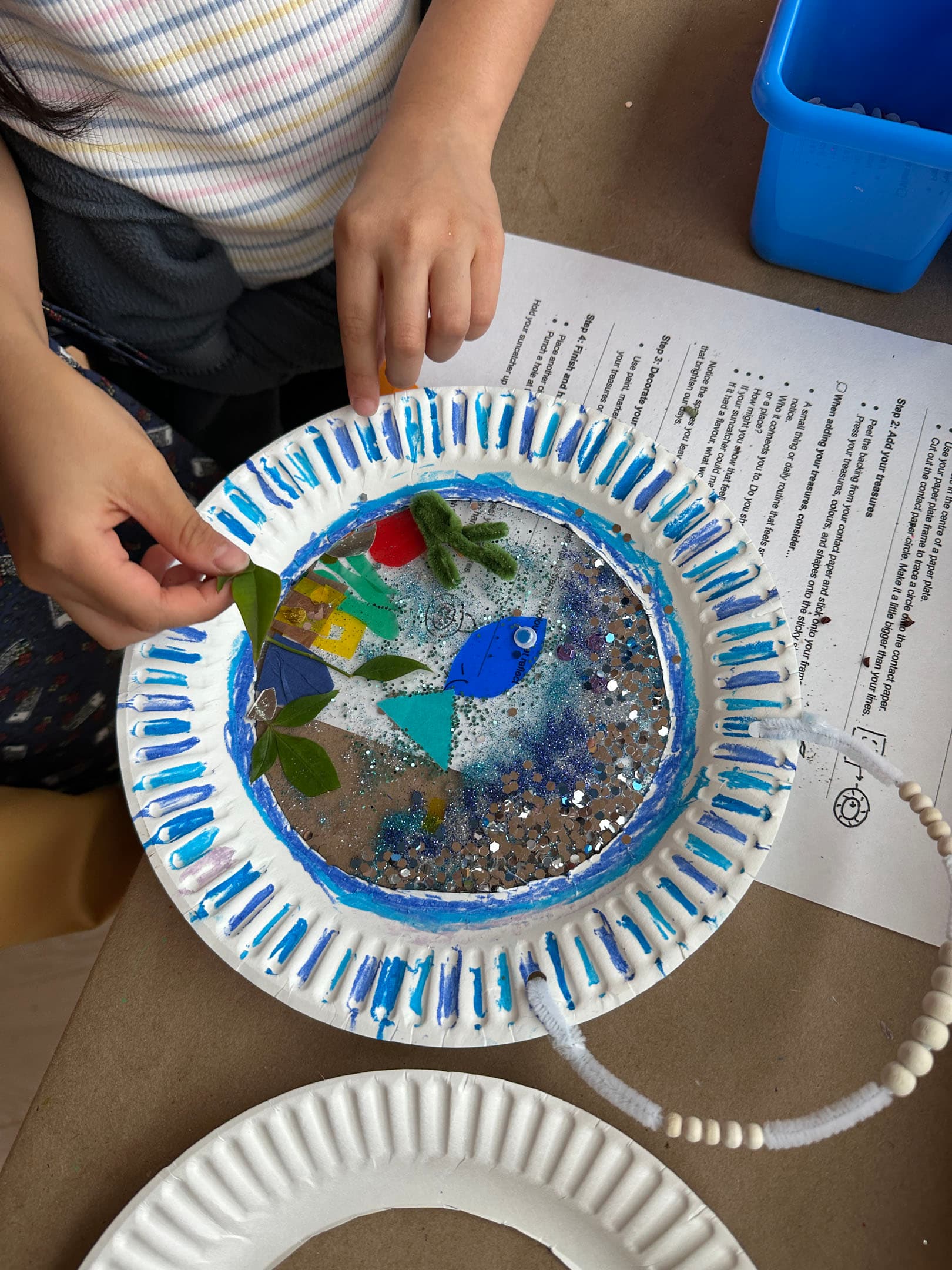 A child's hands are seen working on a decorated paper plate, which features a mix of glitter, colorful shapes, and green leaves. The plate has a blue painted edge and contains a fish illustration among other decorative elements. In the background, there are additional paper plates and instructional sheets on a table.
