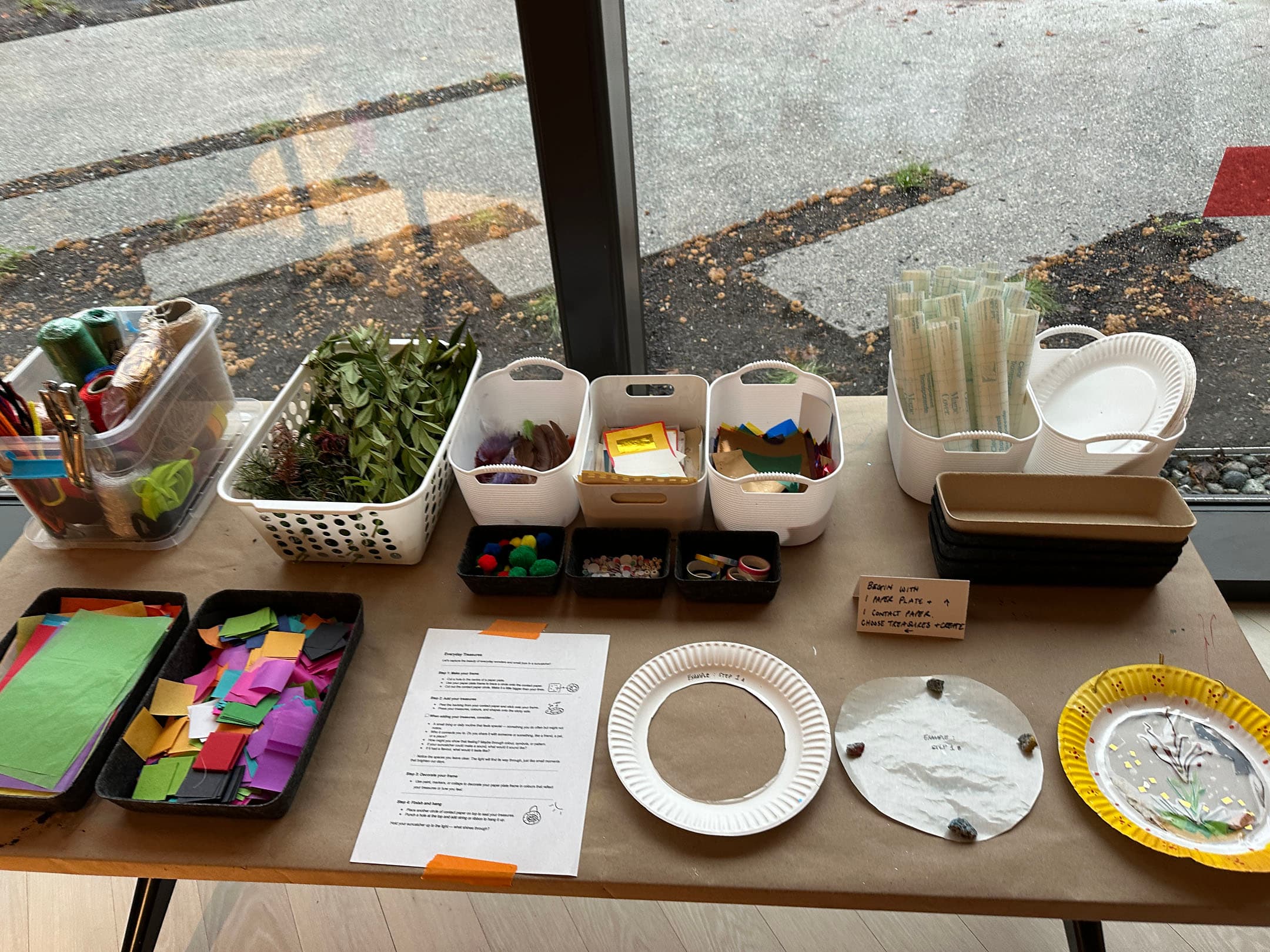 A table displays various crafting supplies and project instructions. On the left, there are containers filled with colored papers, ribbons, scissors, and threads. A basket holds natural materials like leaves and pinecones. Other containers include pom-poms, stickers, and paper plates. Several instructions outline crafting steps. The background shows a window with an outdoor view.