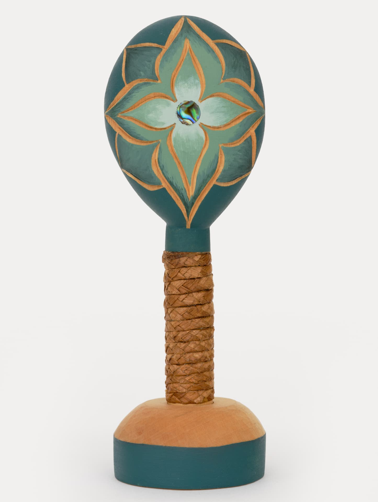 A decorative wooden maraca with a rounded, teal-painted head featuring a floral design in shades of green and gold. The center has a small circular inlay that reflects light. The handle is wrapped in braided natural fibers and is attached to a circular base that has a smooth, natural wood finish, accented with a teal bottom edge. The background is plain and white.