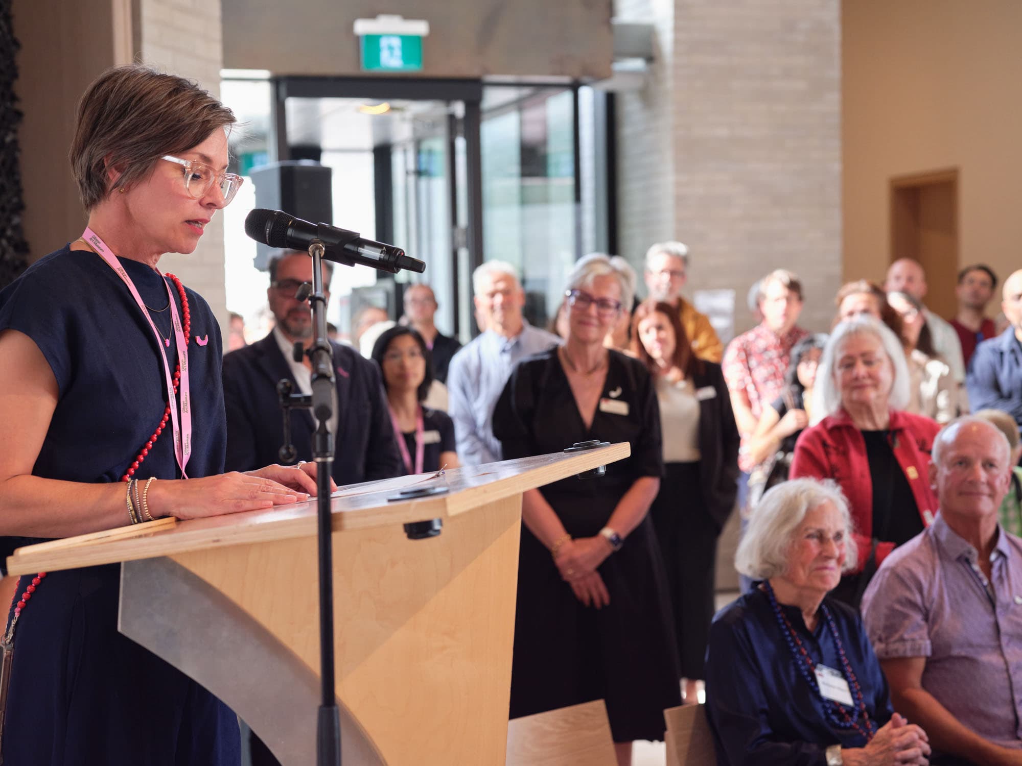 A woman with short hair and glasses stands at a wooden podium, speaking into a microphone. She wears a navy dress with a red necklace and a pink lanyard. In the background, a diverse audience of people listens attentively, some seated and others standing, in a well-lit indoor space. Large glass doors are visible behind the speaker, allowing natural light to fill the room.