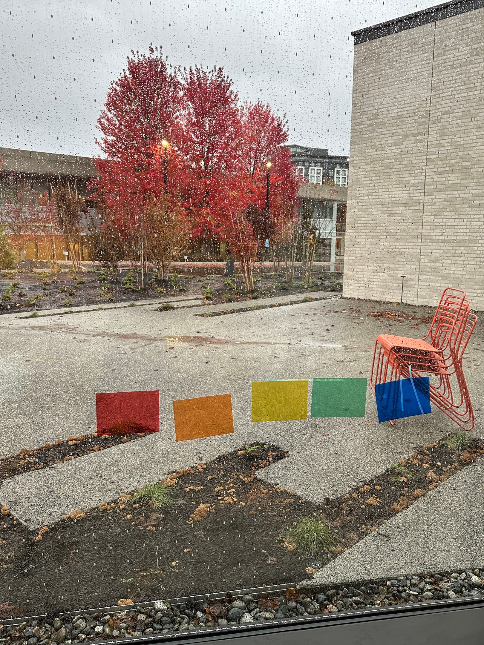A view through a rain-speckled window showing a colorful array of square transparent panels (red, orange, yellow, green, and blue) against a gray pavement. Outside, vibrant red trees are visible, along with a modern building and an orange metal chair placed nearby on the ground. The scene is rainy and overcast.