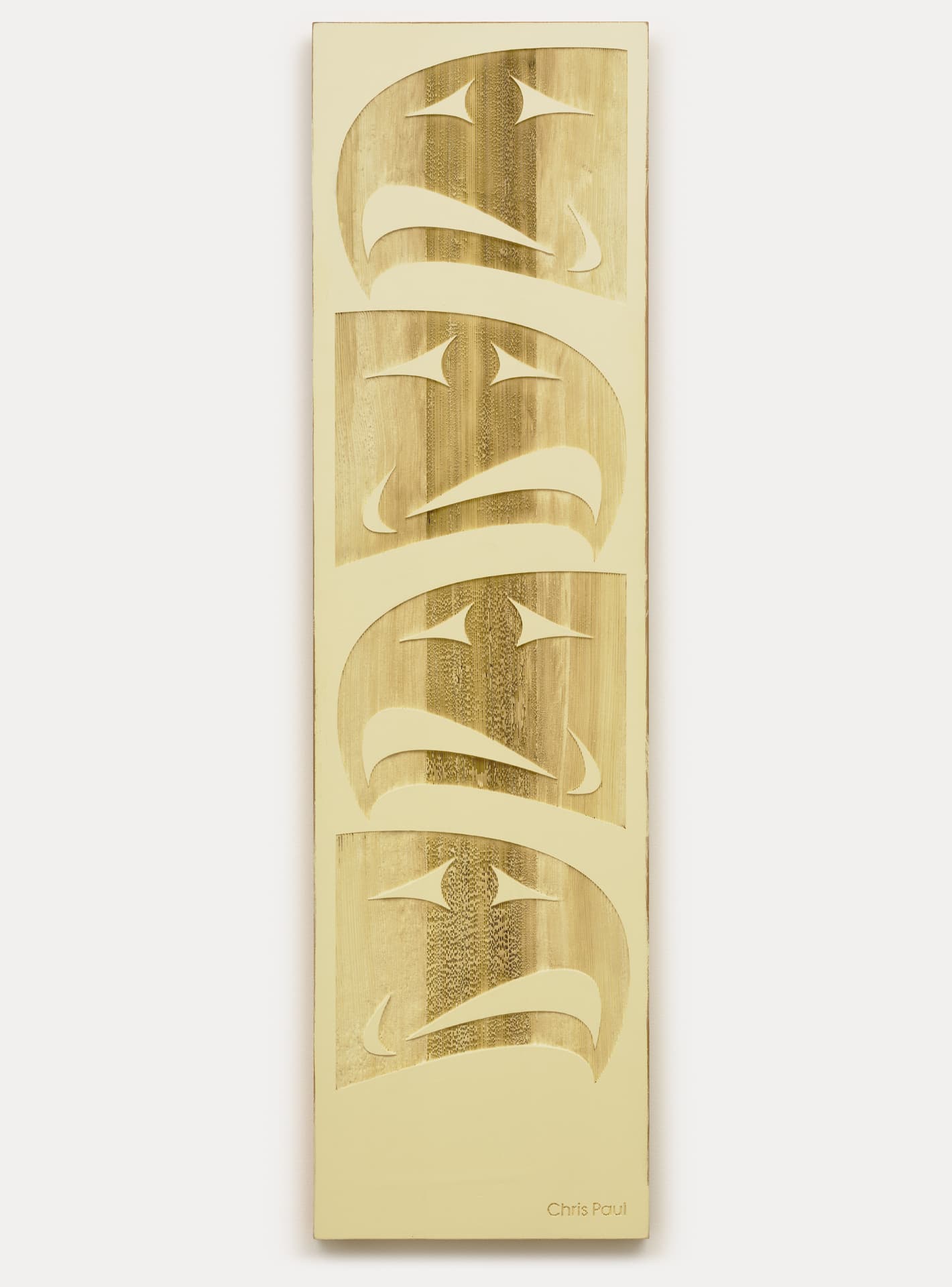 A vertical wooden panel features a series of four stylized, cut-out faces with curved shapes and angular eyes, arranged in a stacked formation. The background has a light golden hue, while the faces have a contrasting darker wood texture. The artist's name, "Chris Paul," is engraved near the bottom.