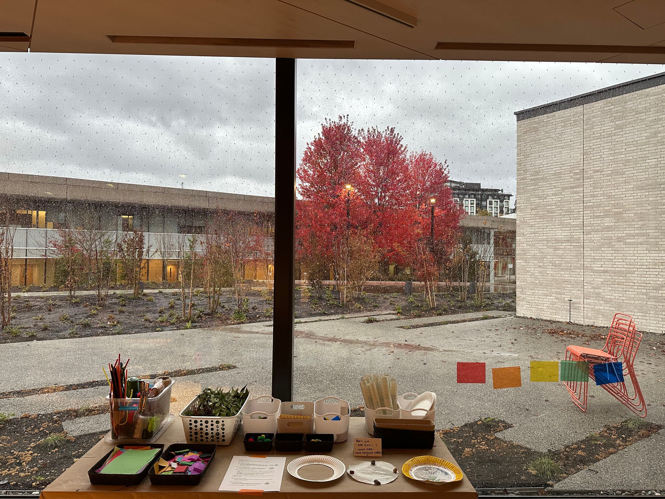 A view from indoors looking out a large window. In the foreground, a table displays various art supplies, including colorful paper, markers, and plates. Outside, a gray, rainy sky looms over a garden with bare trees and a vibrant red tree in the center. To the right, an orange metal chair is positioned on a gray paved area. The scene captures a blend of indoor activities and an autumn landscape.