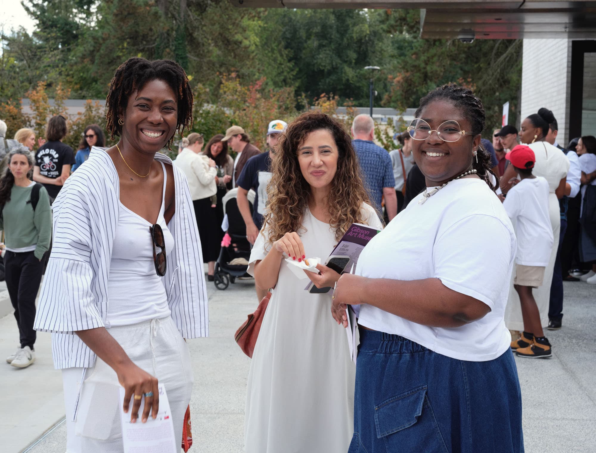 Three women are smiling at the camera in a lively outdoor setting. The woman on the left has dark, curly hair and is wearing a striped cardigan over a white top. The woman in the middle has long, curly hair and is dressed in a light-colored dress. The woman on the right is wearing glasses and a white shirt paired with a denim skirt. In the background, several people are gathered, creating a festive atmosphere.
