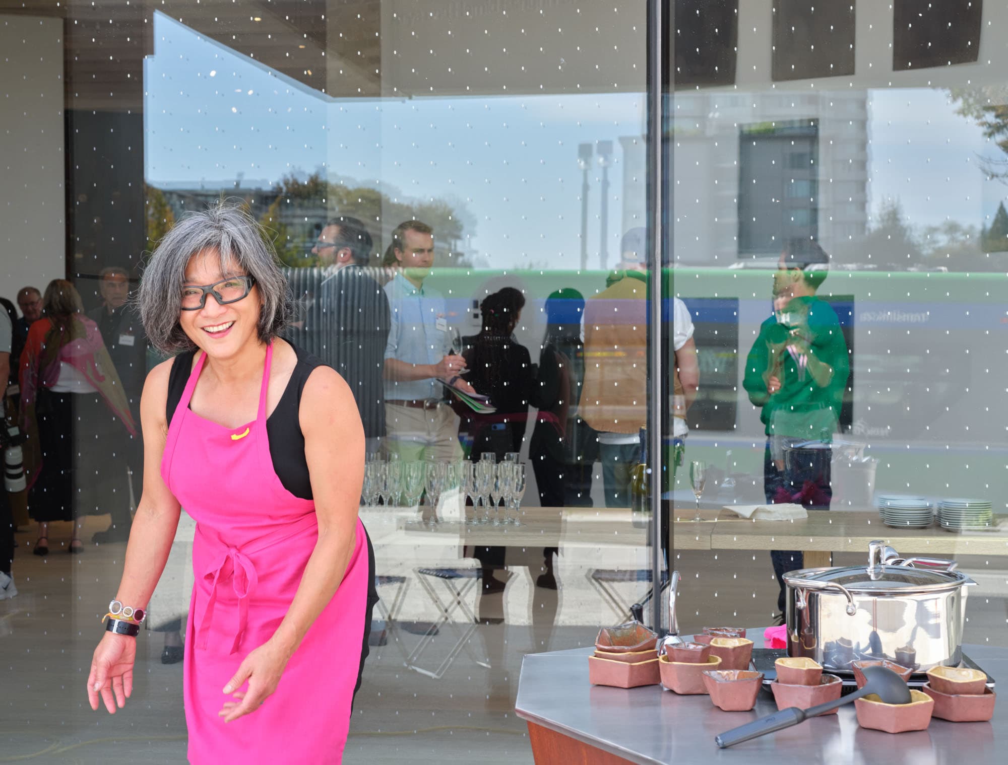 A smiling woman with short, gray hair and glasses wears a bright pink apron while standing outside a glass window. In front of her is a table with several food containers and a pot. Reflected in the glass are people mingling in the background, some wearing name tags, with a modern building visible. The setting appears to be an outdoor event or gathering.