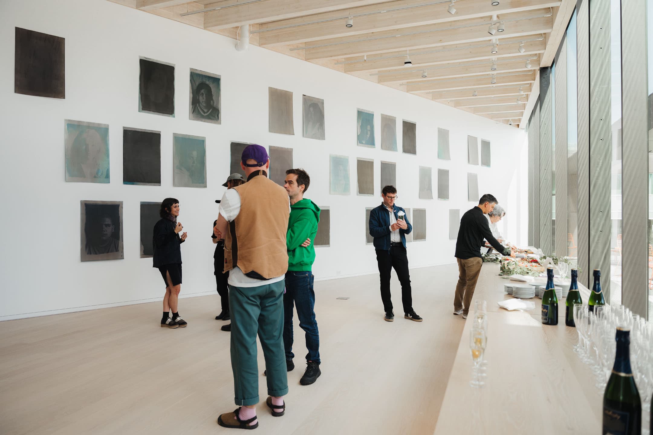 A spacious gallery features a collection of large, muted portraits on the wall. In the foreground, a group of people are engaged in conversation, while one individual stands apart, inspecting a phone. A long table along the side holds food and drinks, including champagne bottles and glasses, illuminated by natural light from large windows. The interior has a modern design with wooden beams and light-colored flooring.