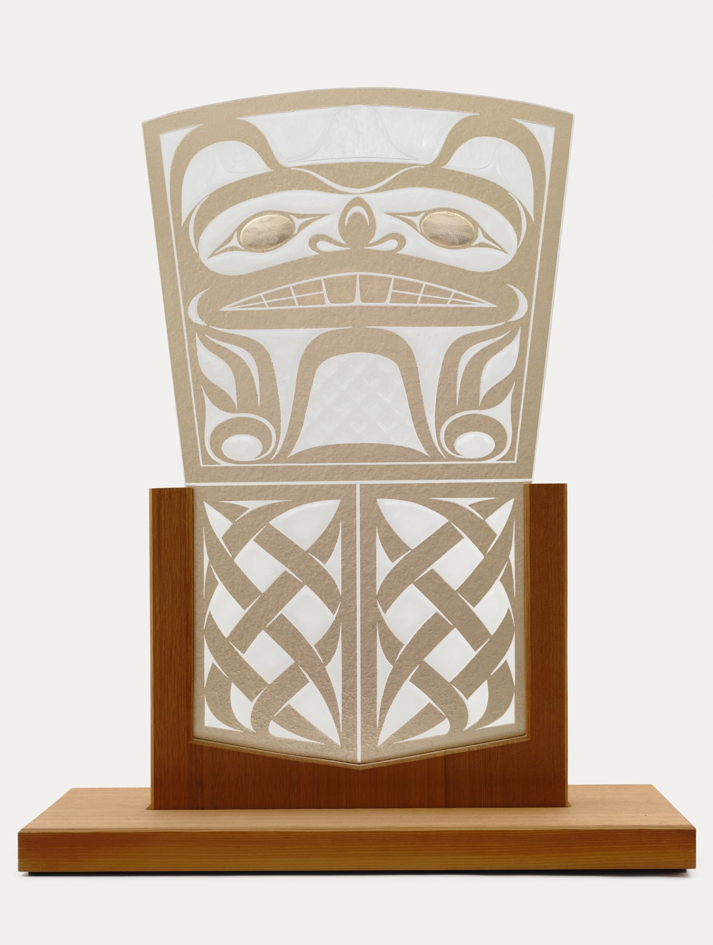 A decorative art piece featuring a stylized mask or face design, crafted from layered materials. The top portion depicts a mask with a prominent expression, characterized by bold outlines and two golden eyes. Below, there is a geometric pattern made of intertwined shapes, all mounted on a wooden base. The overall color scheme includes cream, beige, and brown, set against a white background.