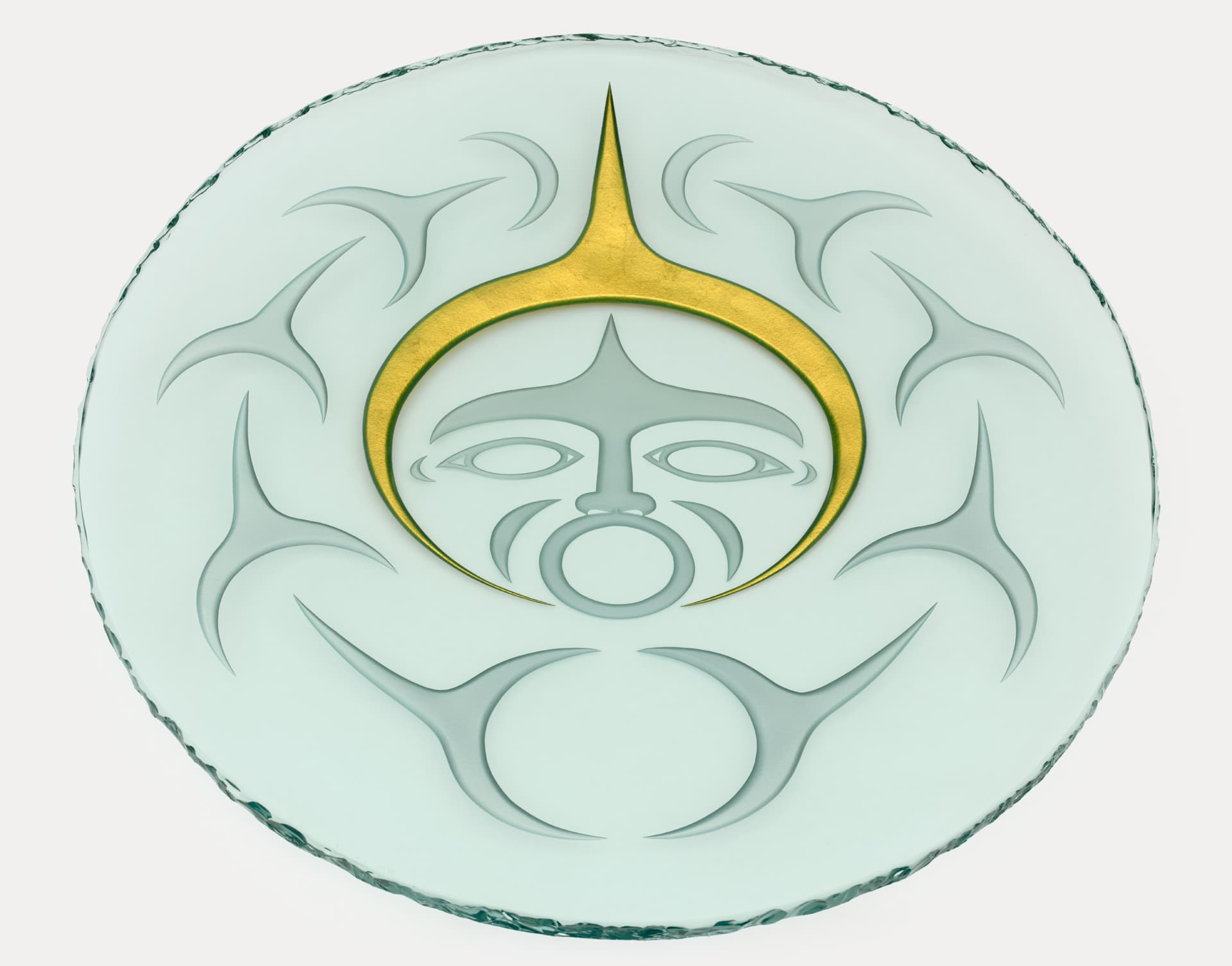 A circular glass plate featuring an embossed design. In the center, there is a stylized face with a prominent golden crescent above it, surrounded by several abstract, curved shapes in a light, frosted finish. The edge of the plate is uneven and textured.
