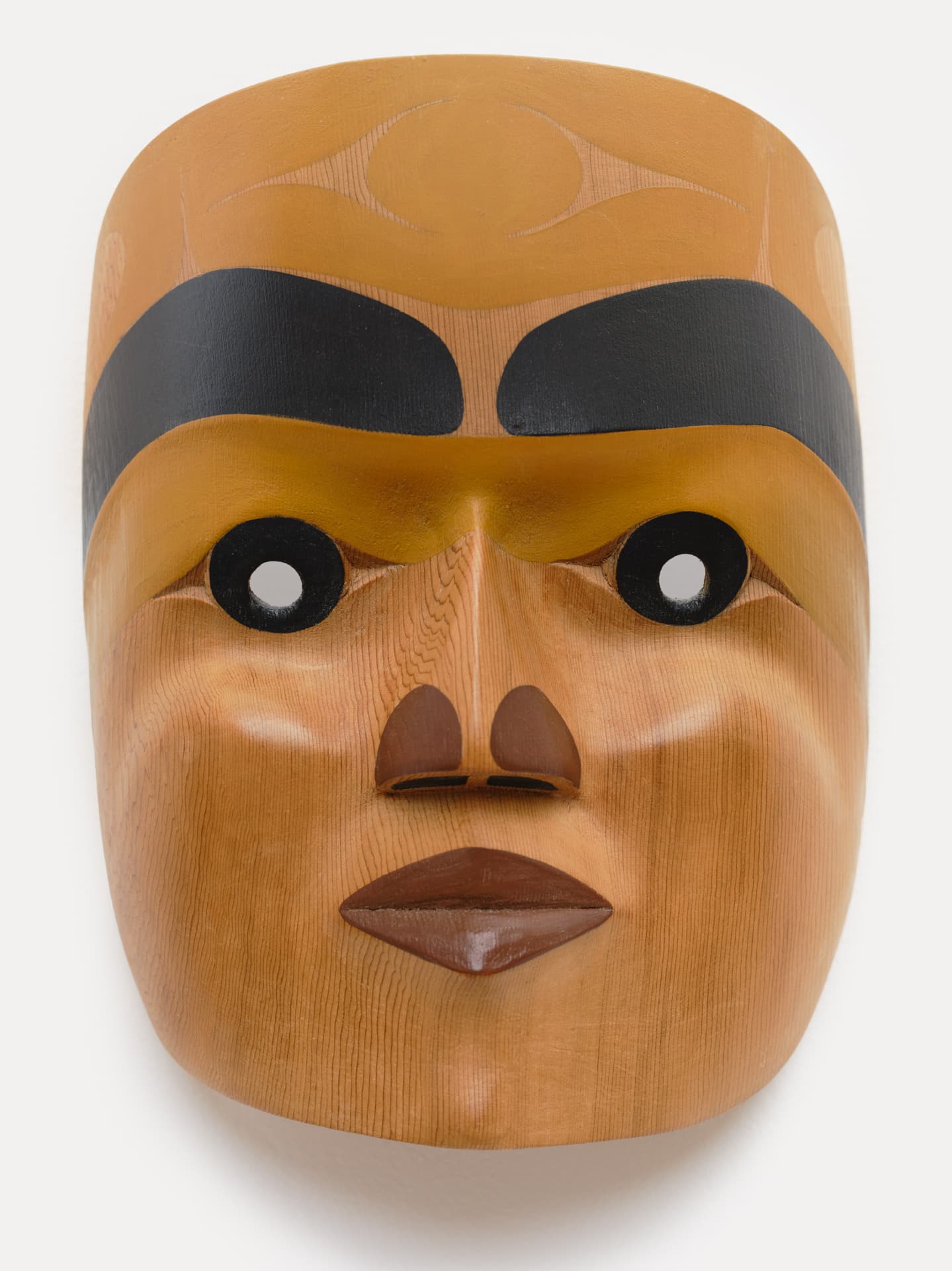 A wooden mask with a rounded shape, featuring a smooth, light-toned surface. The mask has prominent black eyebrows and large, circular eyes with white pupils. The nose is stylized and slightly raised, while the lips are plump and painted brown. Subtle wood grain lines are visible throughout the surface. The mask is displayed against a plain white background.