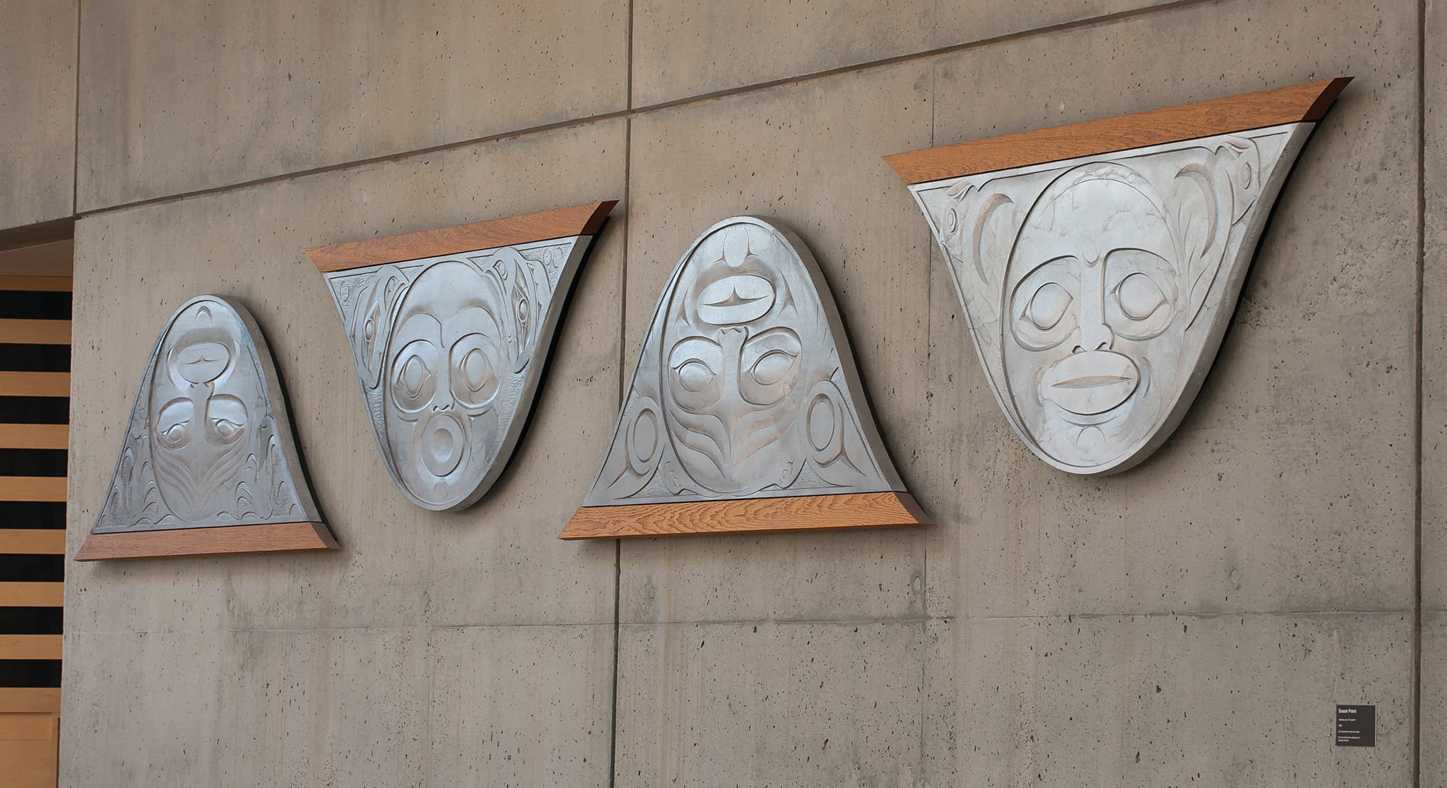 Four stylized metal masks are mounted on a concrete wall, each with a triangular shape and wooden bases. The masks feature exaggerated facial expressions, showcasing distinct eyes, mouths, and decorative patterns. The background is plain and textured, highlighting the artistic elements of the masks. A small plaque is visible to the right.