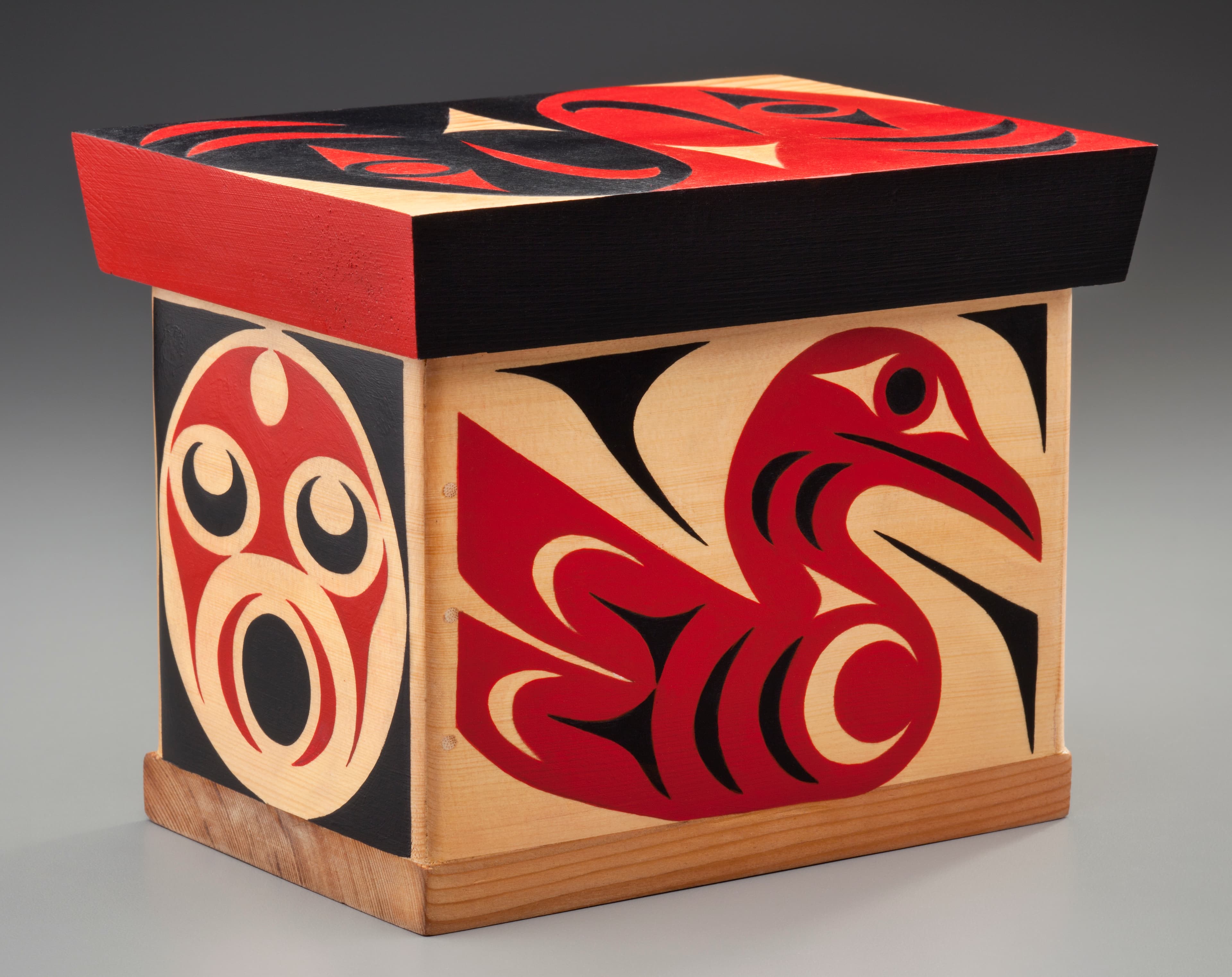 A wooden box with a vibrant design, featuring a red and black color scheme. One side depicts a stylized red bird with black outlines, while another side shows a circular design with facial features in red and black. The box has a smooth, light-colored wood finish and a solid red top. The overall design reflects artistic patterns characteristic of indigenous art styles.