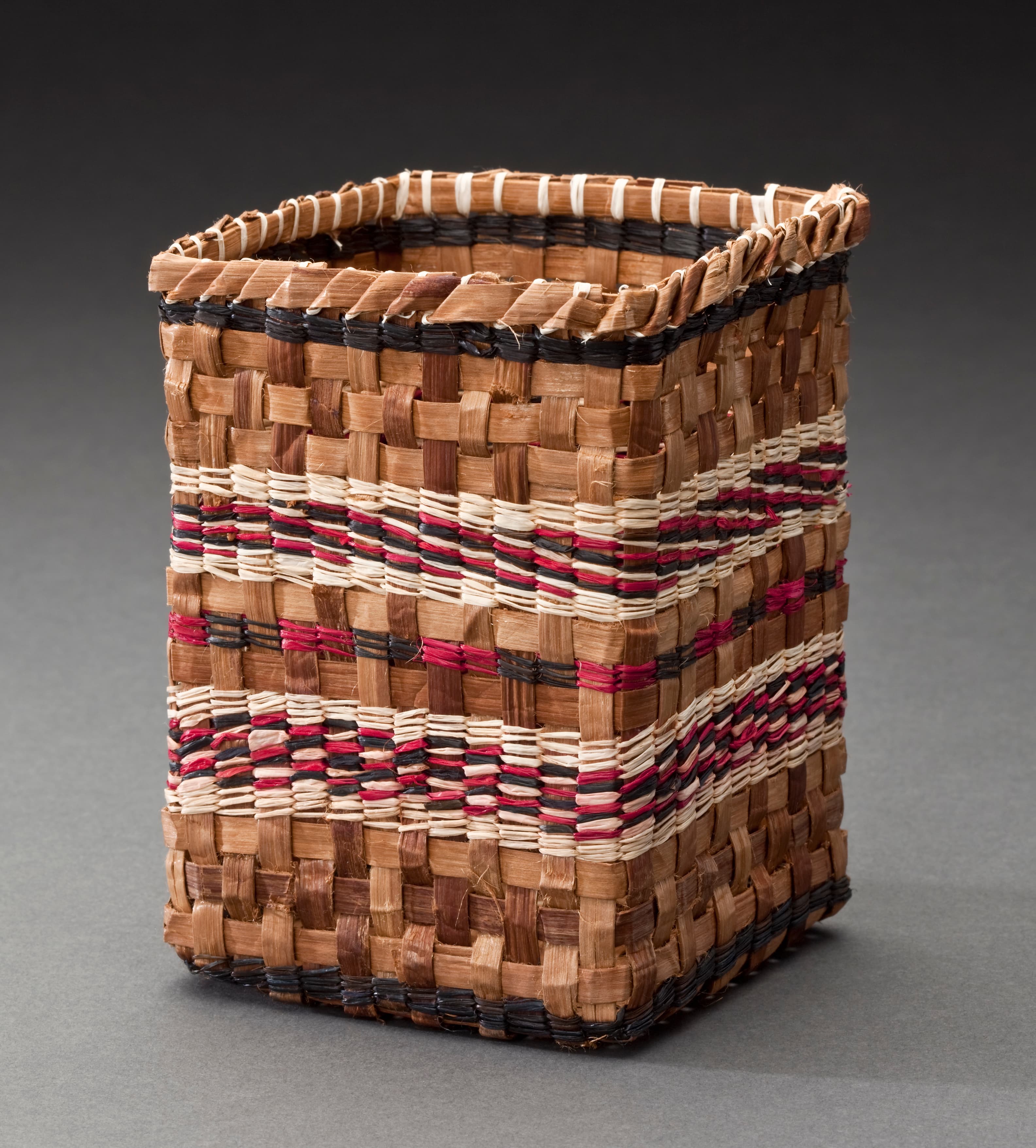 A woven rectangular basket made from natural fibers in shades of brown, cream, and black, with decorative patterns featuring red stripes. The basket has an open top and a structured, straight-sided design. It is set against a gray background.