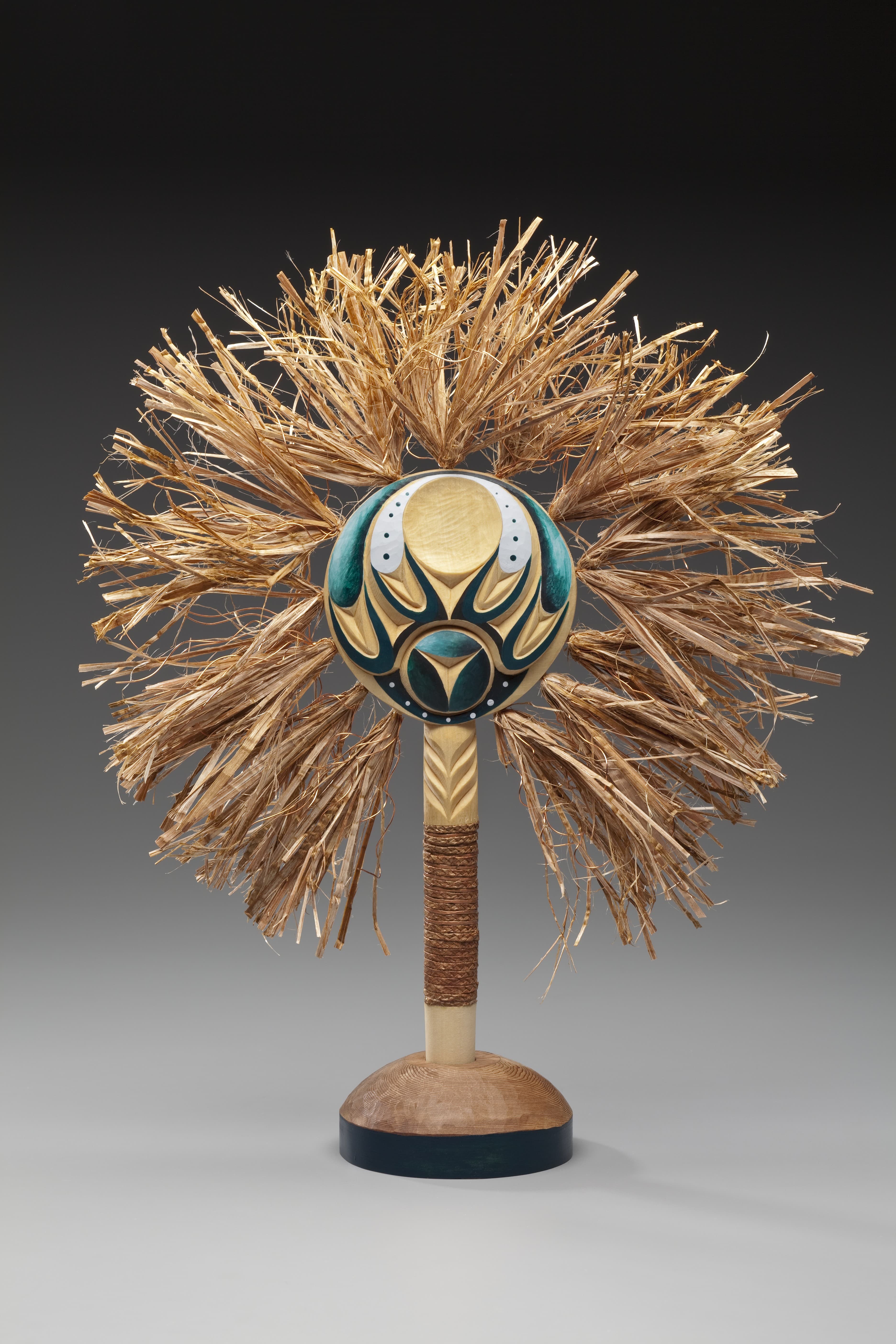 A decorative object featuring a rounded base made of wood and a tall, slender pole wrapped in natural fibers. At the top, a vibrant circular element is adorned with blue and gold patterns, surrounded by a fan of dried grass-like strands arranged to resemble hair or foliage. The background is a gradient from light to dark gray, emphasizing the sculpture's intricate design.