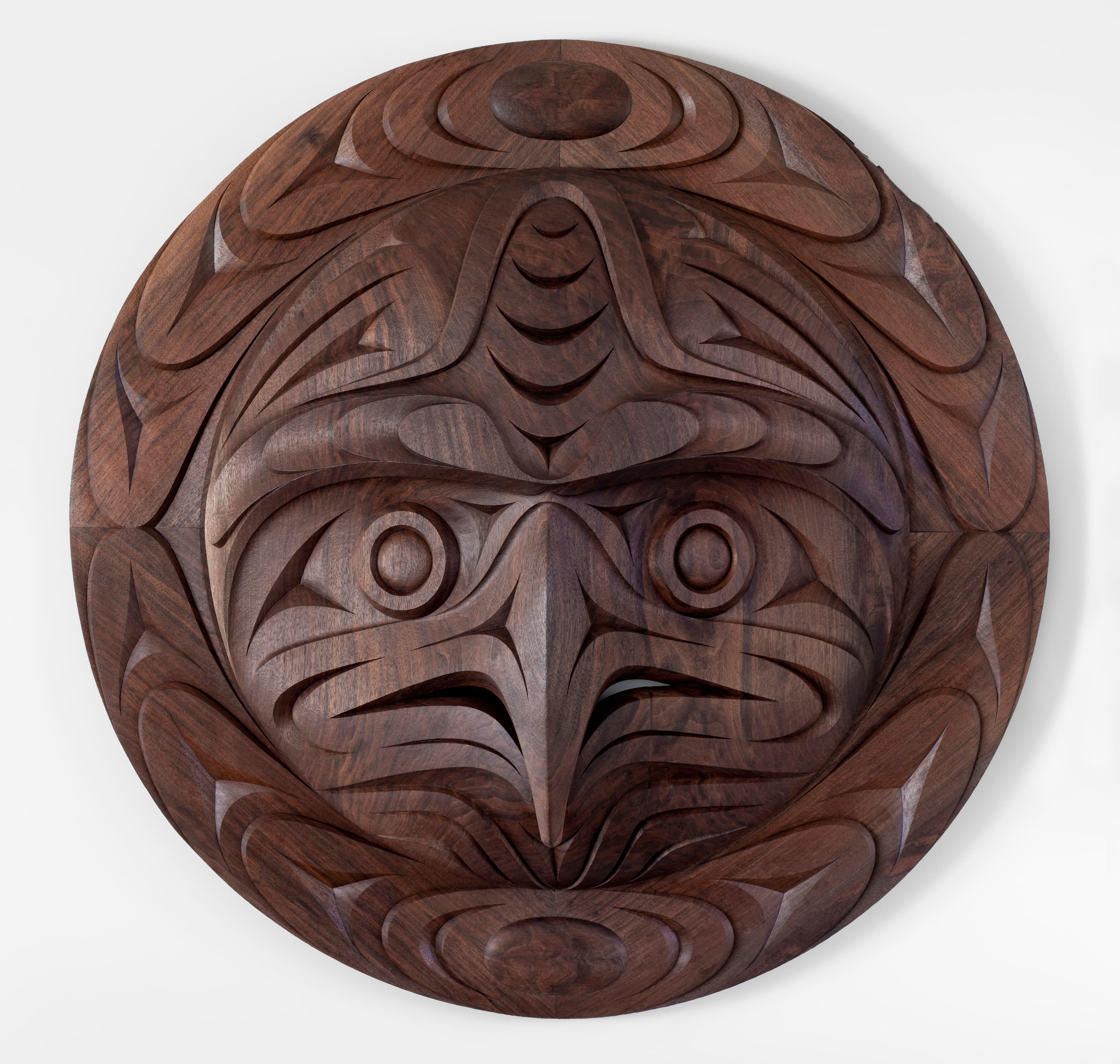 A circular wooden plaque features intricate carvings of a stylized bird face, displaying prominent eyes and a sharply defined beak. The wood appears to be a rich, dark color, enhancing the depth of the carvings that include abstract patterns surrounding the central design. The background is plain, allowing the artwork to stand out.