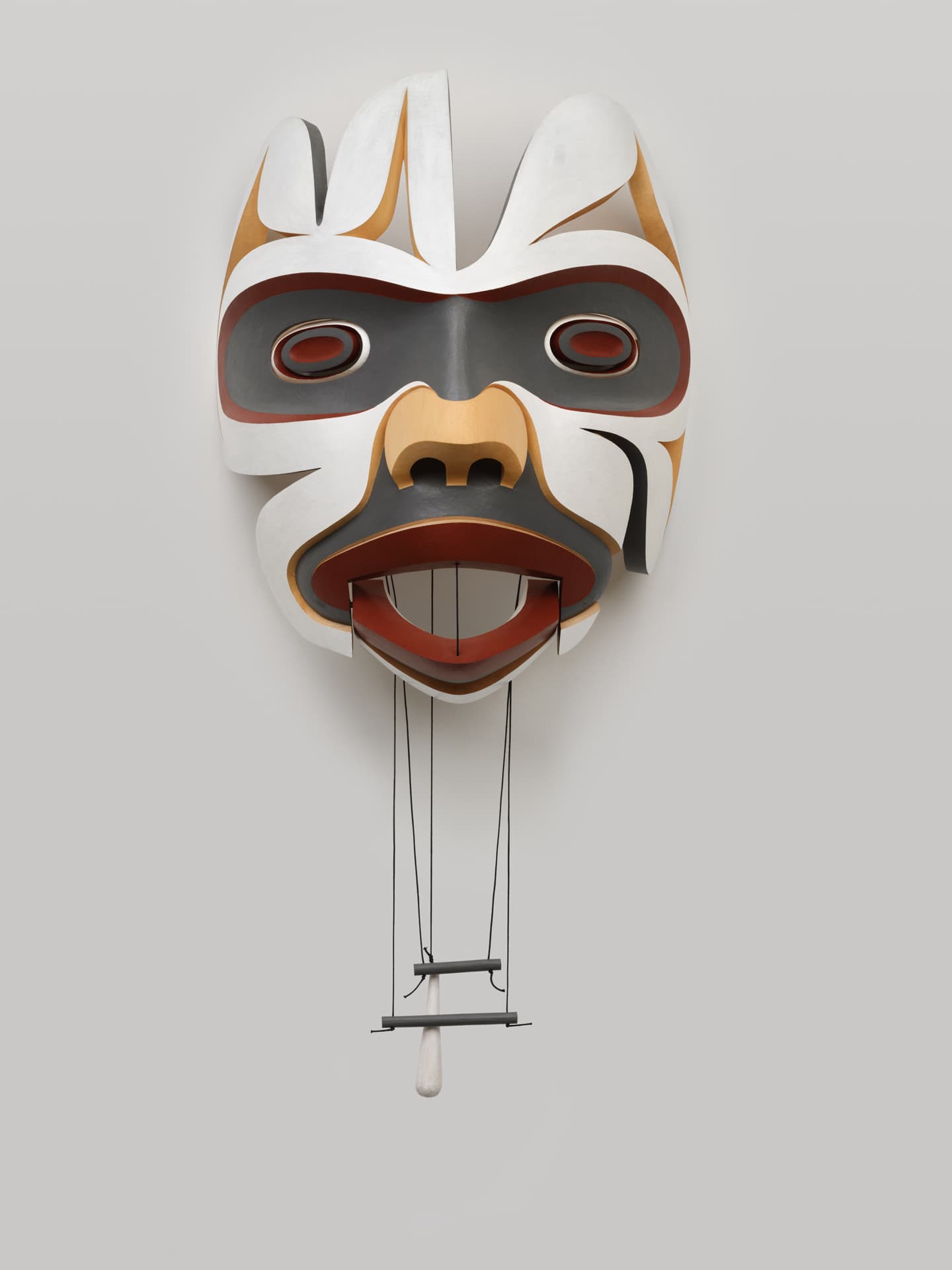 A large, stylized mask hangs against a plain light gray wall. The mask features a combination of white, black, red, and brown colors, with exaggerated facial features, including large eyes, a broad mouth, and intricate patterns. Below the mask, there are black strings leading to a small wooden element suspended by a horizontal bar. The overall composition emphasizes the mask's bold design and vivid colors.