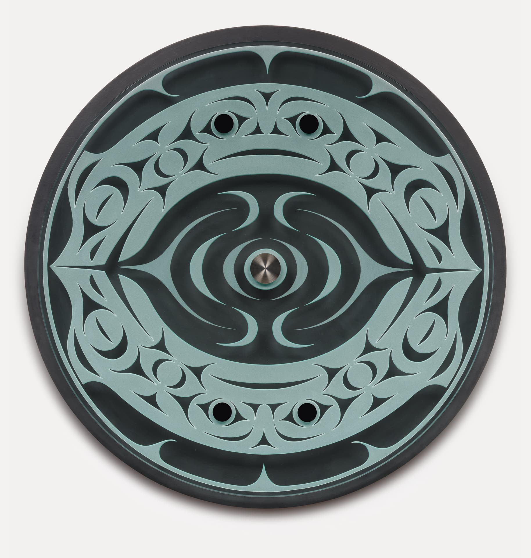 A round, decorative object featuring intricate teal and black patterns. The design includes swirls and sharp shapes radiating from a central metal circle. The outer edge is smooth and black, enhancing the contrast of the detailed design on the surface. Two small holes are positioned symmetrically within the pattern.