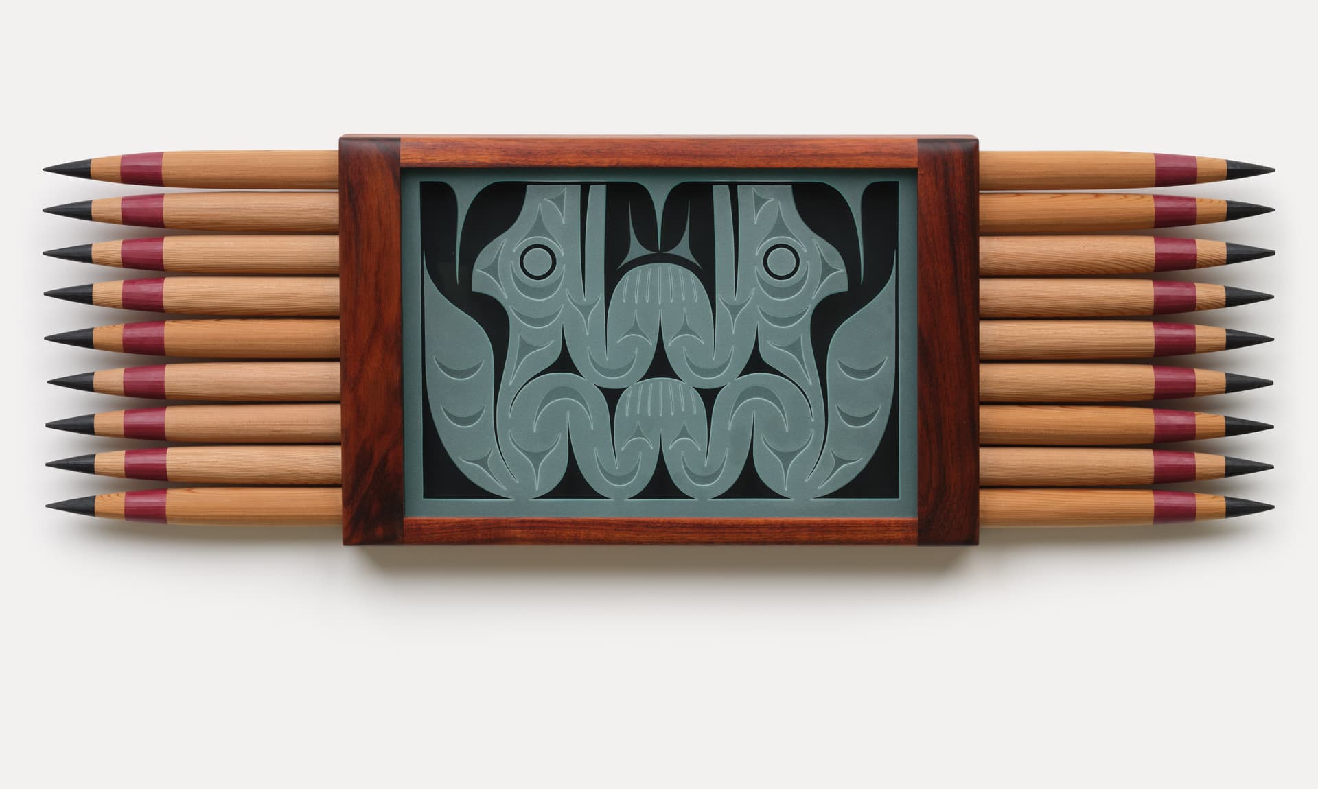A collection of wooden pencils arranged in two rows, with a decorative central piece featuring embossed abstract designs. The pencils have pointed tips, with some colored in a reddish hue, framed by a dark wooden casing around the decorative insert. The background is plain white, emphasizing the colorful and textured elements.
