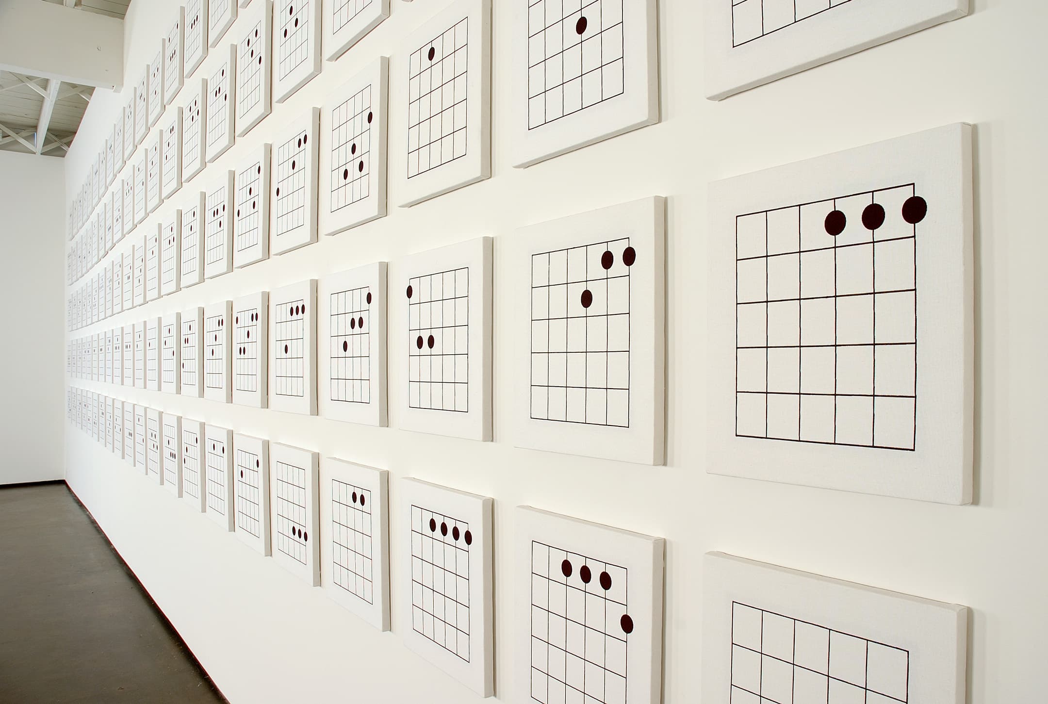 A wall displays a grid of framed artworks, each featuring a diagram of guitar chord shapes. The diagrams consist of a grid with black lines and various black dots positioned on the strings, set against a plain white background. The arrangement includes multiple rows and columns of these chord illustrations.