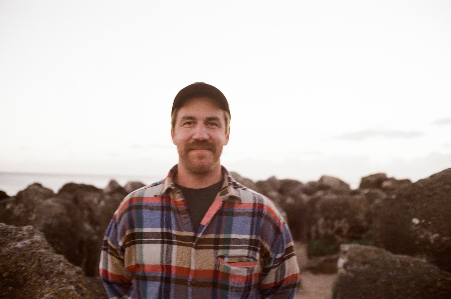 A man with a mustache is standing on a rocky shoreline, wearing a plaid flannel shirt and a black cap. The background features the ocean and a light-colored sky during dusk. The focus is on the man's smiling face, with rocky formations in soft focus around him.