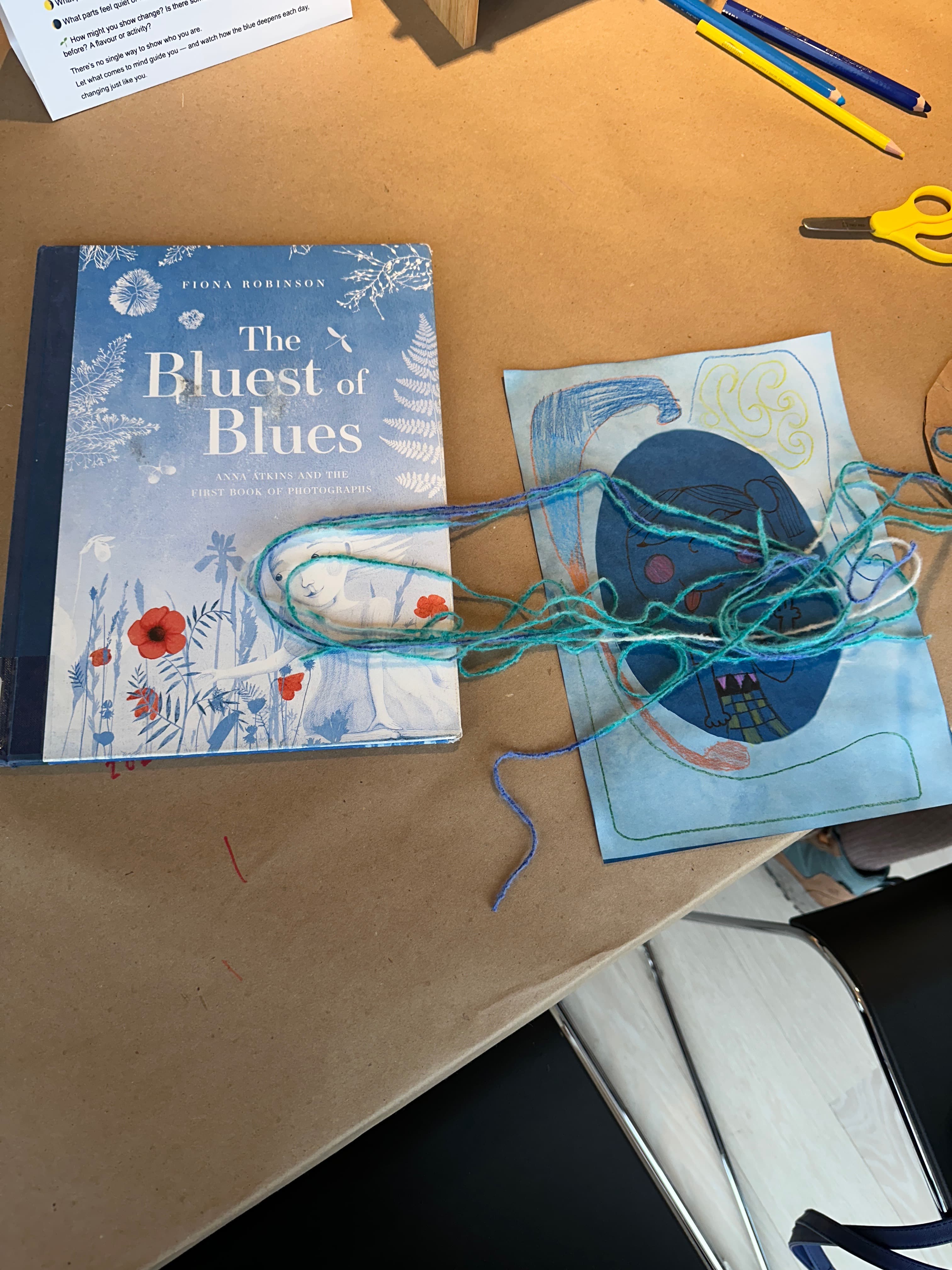 A blue hardcover book titled "The Bluest of Blues" by Fiona Robinson is placed on a brown surface, next to a colorful drawing featuring a blue figure with a smiling face. A piece of multicolored yarn and some art supplies, including scissors and colored pencils, are also visible in the scene.