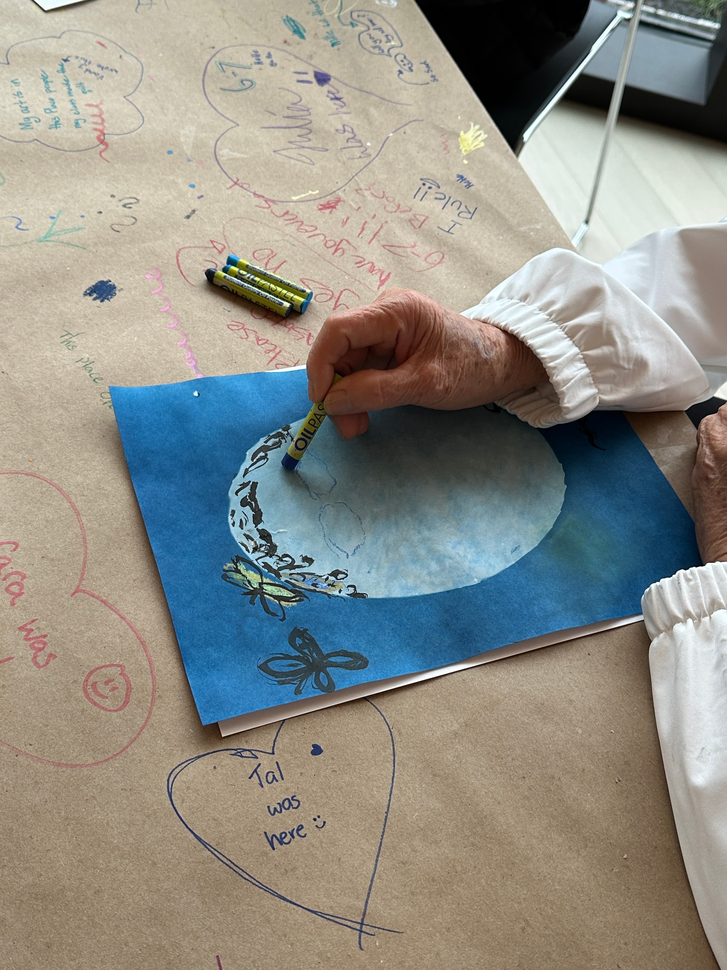A hand is shown holding a yellow crayon, drawing on a blue sheet of paper that depicts a circular, whimsical scenery with butterflies. The background features a table covered in brown paper with various colorful scribbles and messages, including heart shapes and names. Two crayons, one yellow and one blue, are placed on the table beside the drawing.